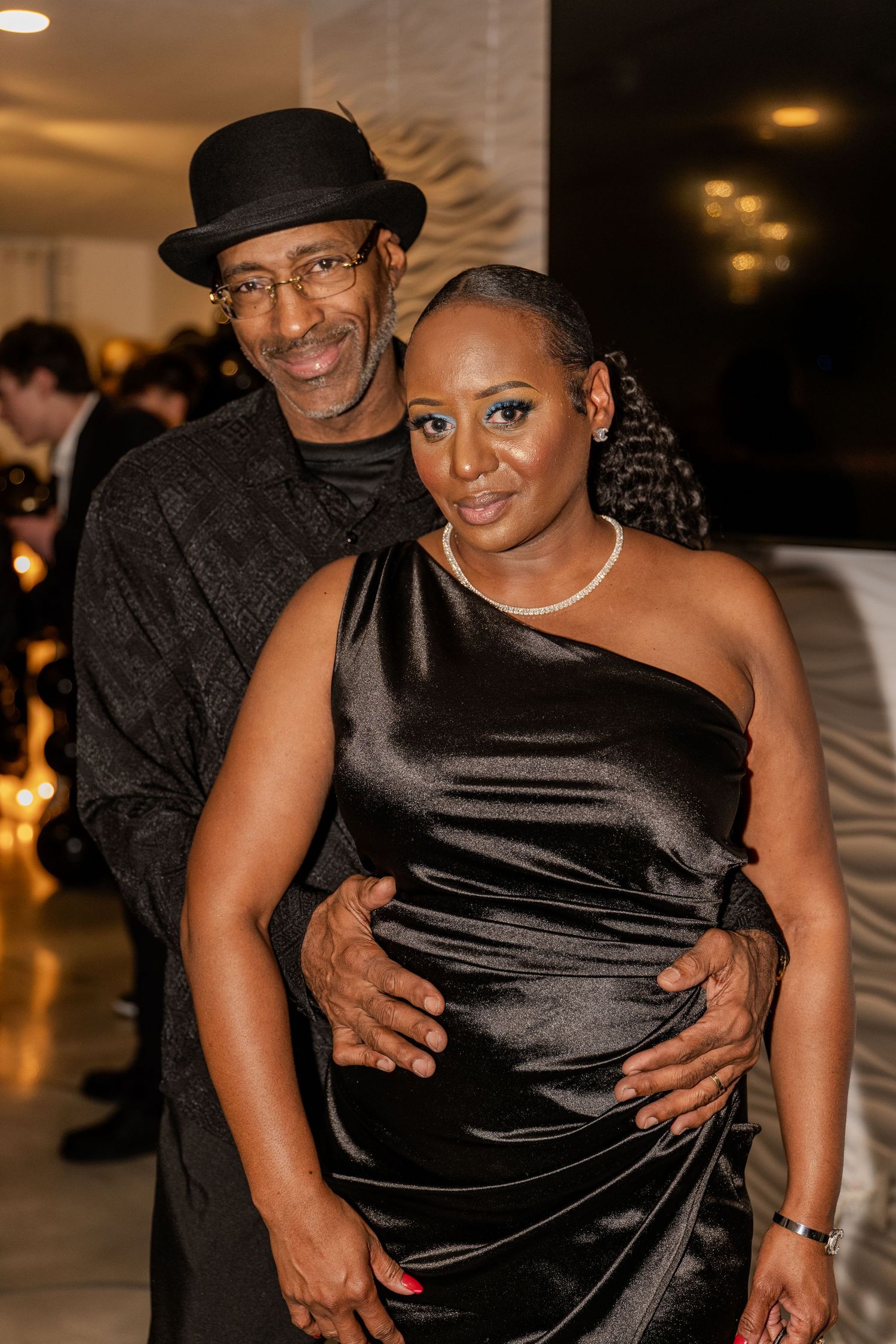 A couple embraces at an elegant evening event, with the person in a black hat and the other in a one-shoulder black satin dress.