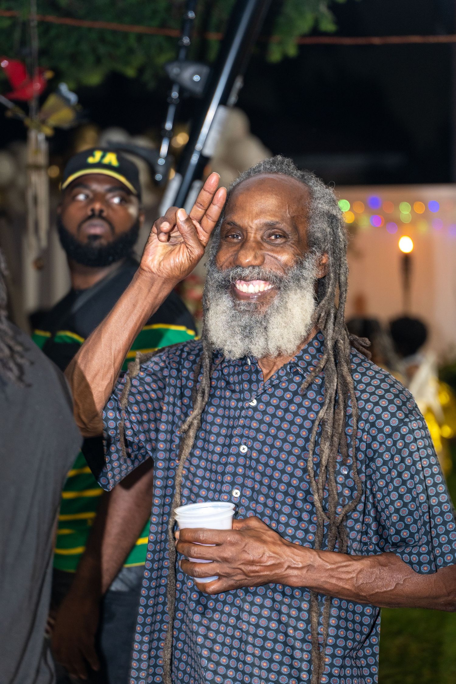 Elderly man with long dreadlocks and grey beard makes peace sign while holding drink.