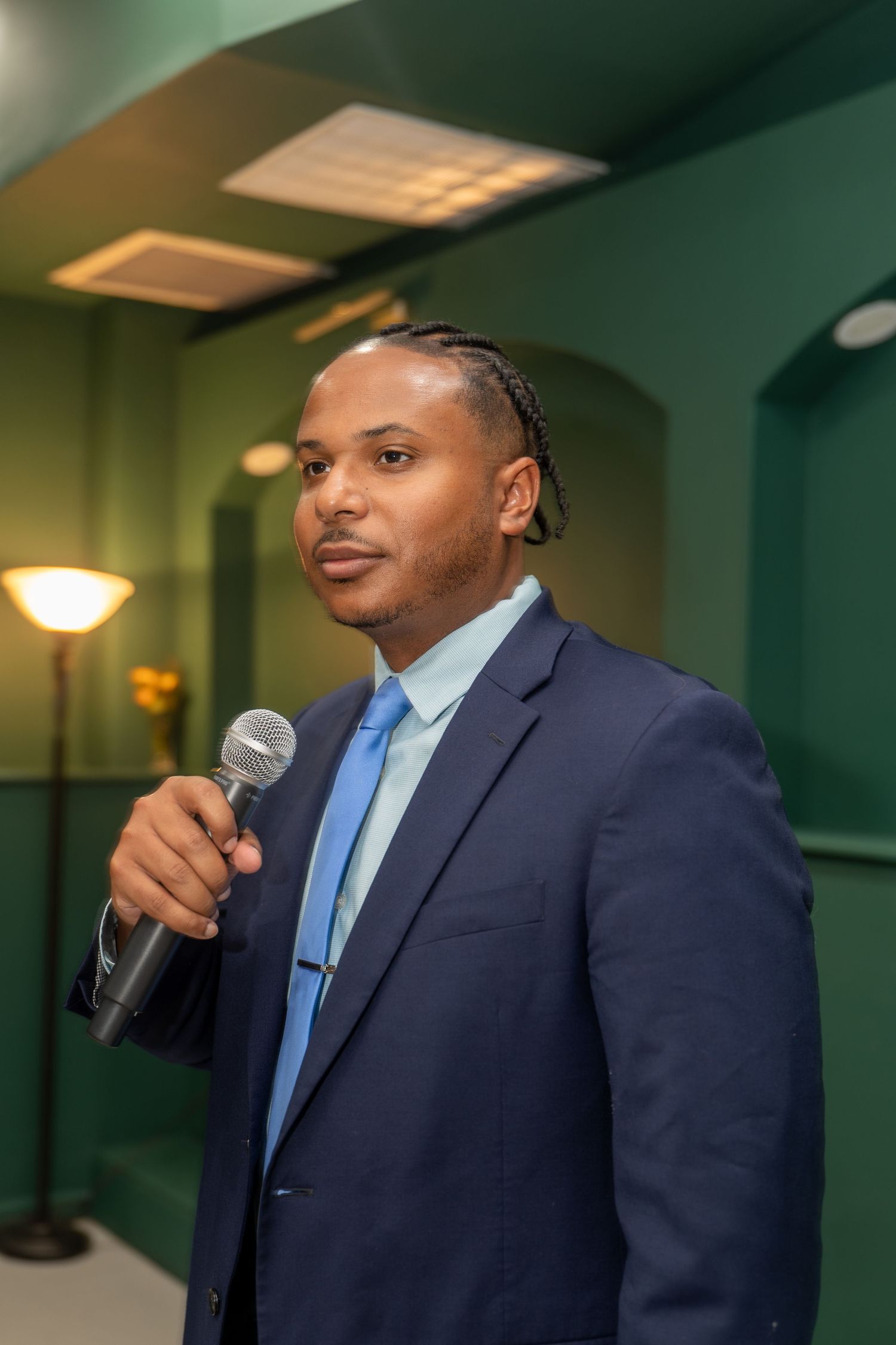 A person in a navy suit and blue tie speaks into a microphone during a presentation.