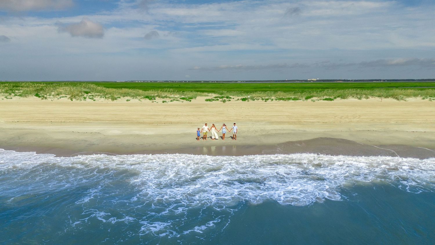 Aerial view of wild horses standing on a sandy beach between ocean waves and green dunes.