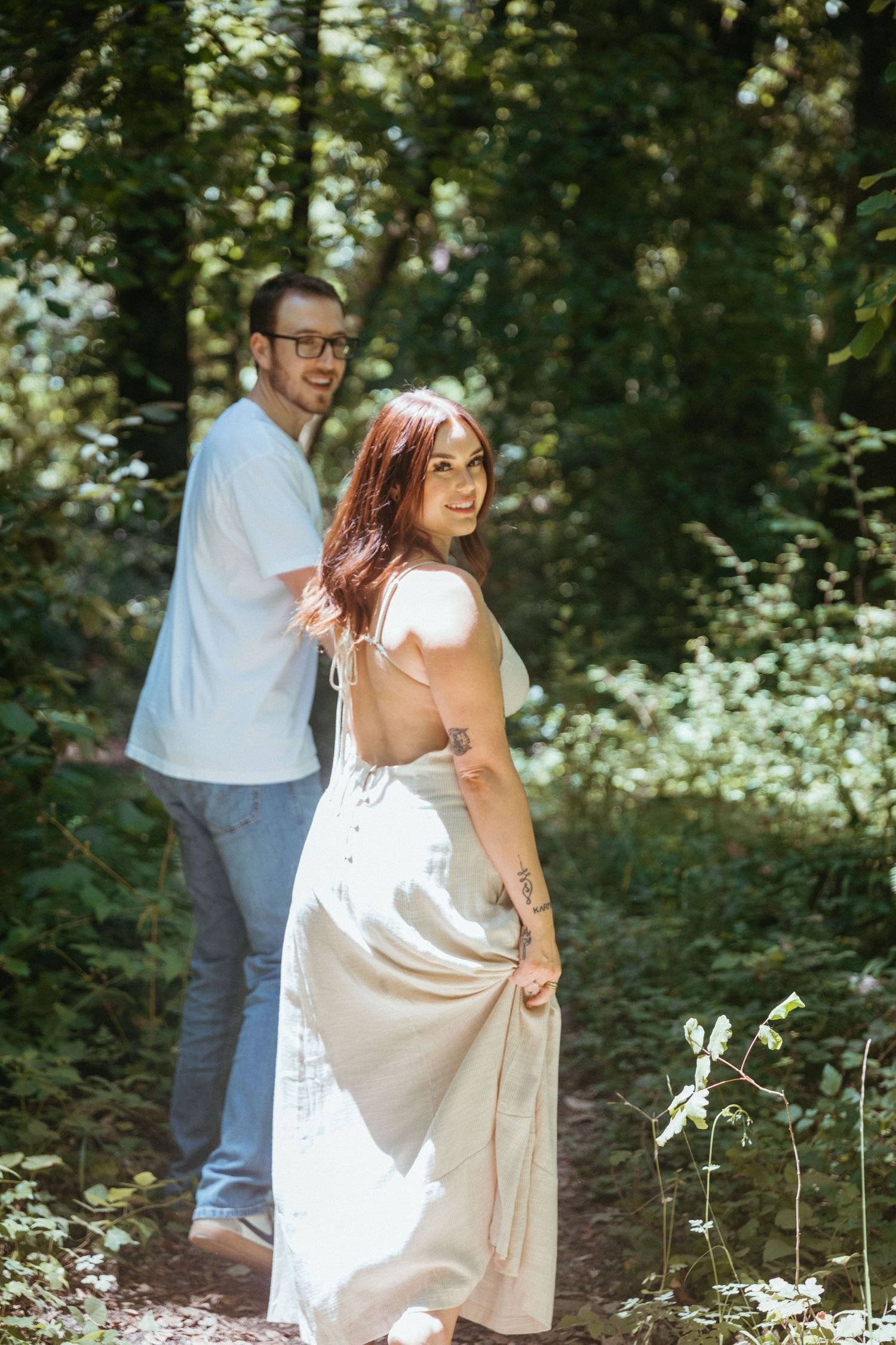 A couple in light-colored clothing walks along a wooded forest path on a sunny day.