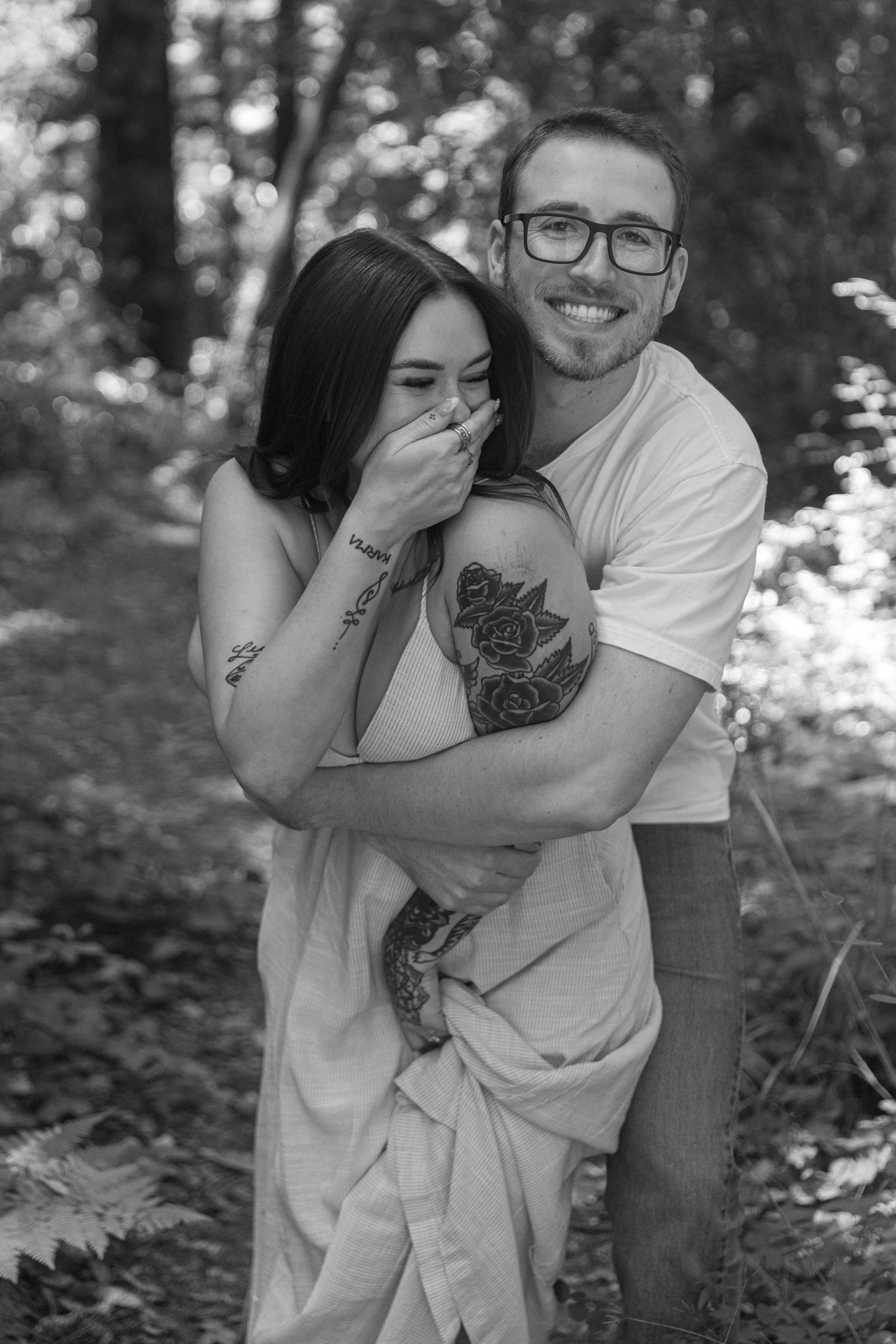 Black and white portrait of embracing couple sharing laughter in forest setting.