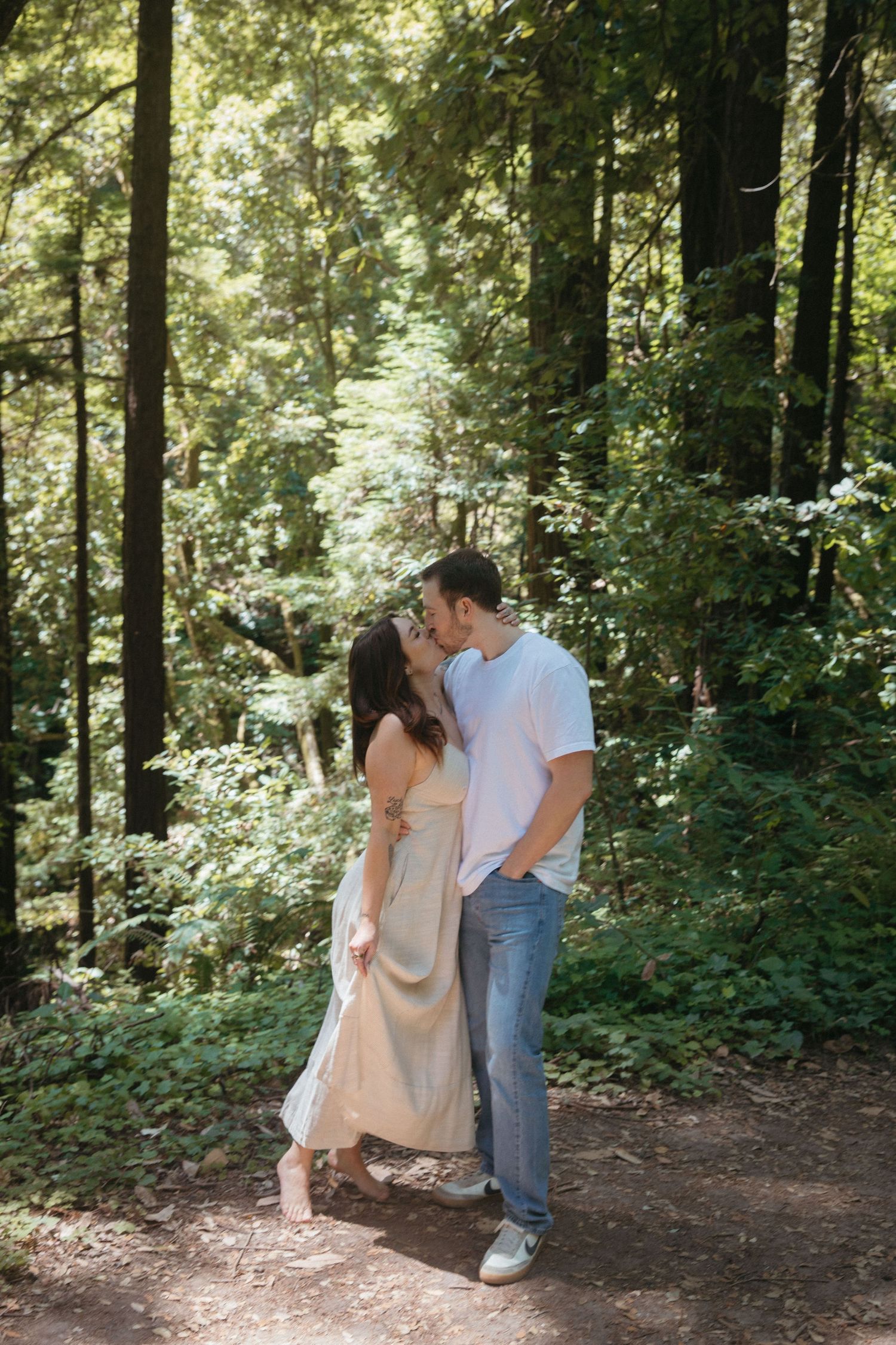 A romantic couple shares an intimate moment on a sunlit forest trail among tall trees.
