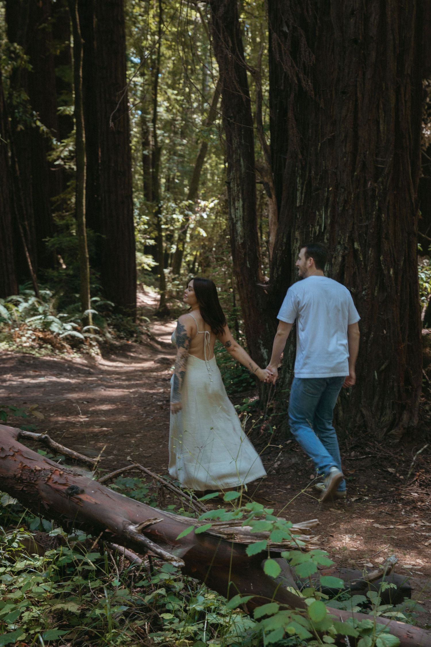 A couple explores a winding forest trail surrounded by towering redwood trees.