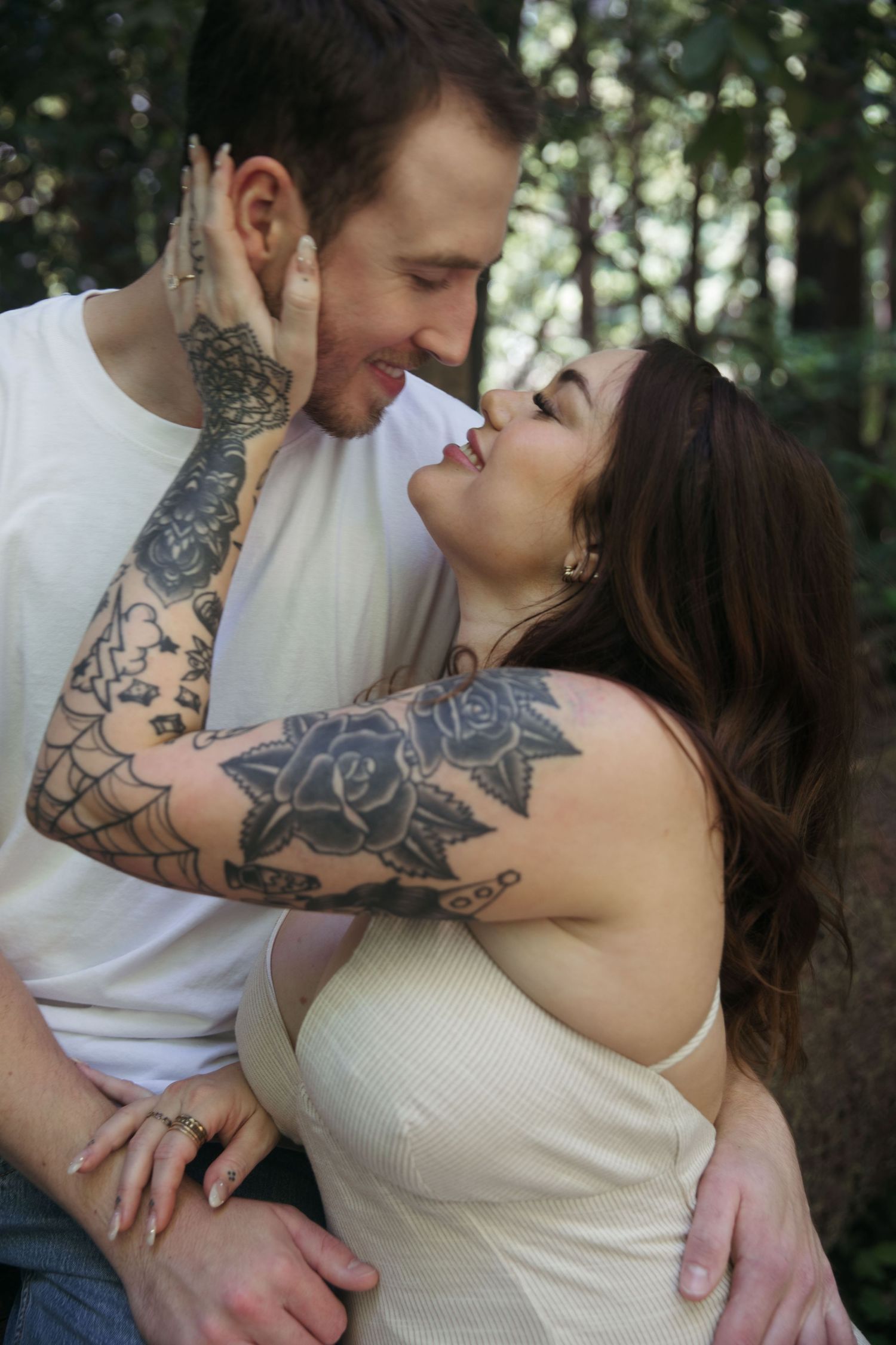 An intimate portrait showing a tattooed couple in a tender embrace surrounded by forest foliage.