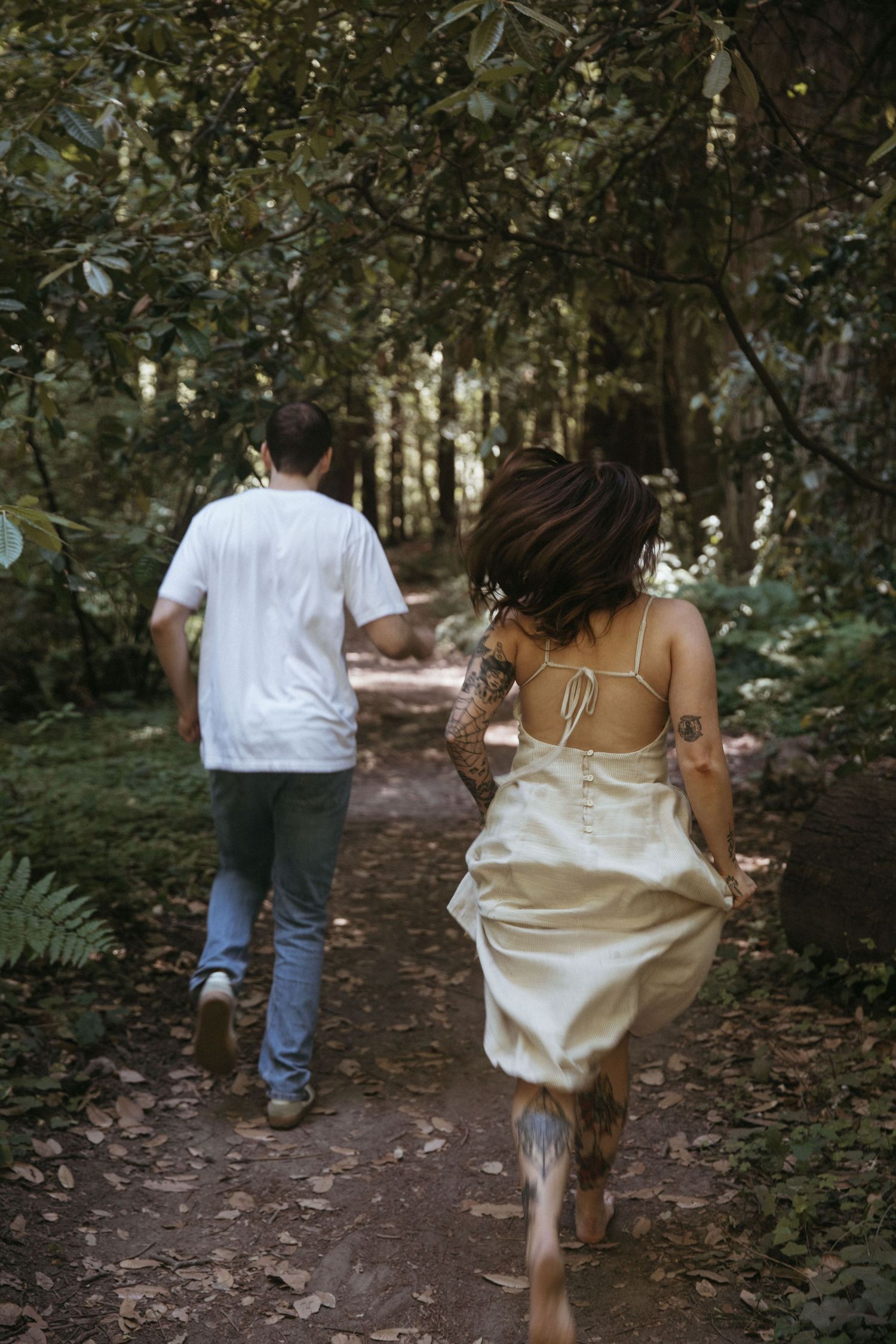 A couple playfully runs down a forest trail with the person in white dress trailing behind.