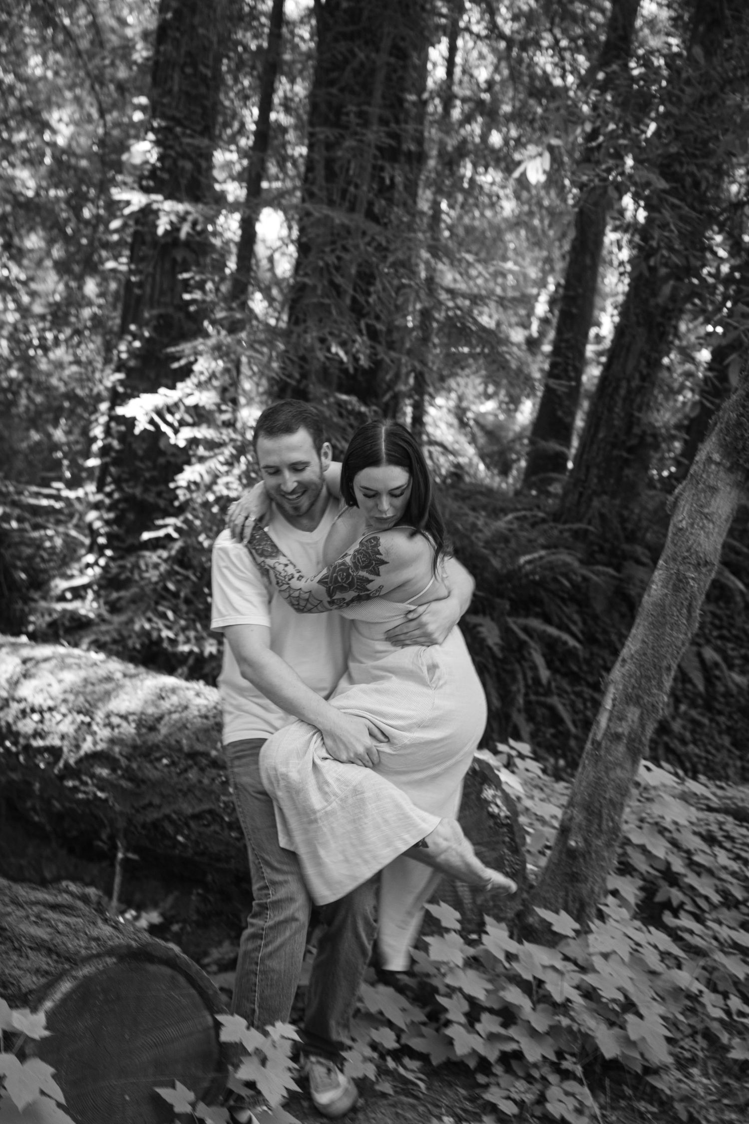 Black and white photo of couple playfully embracing in a wooded forest setting.