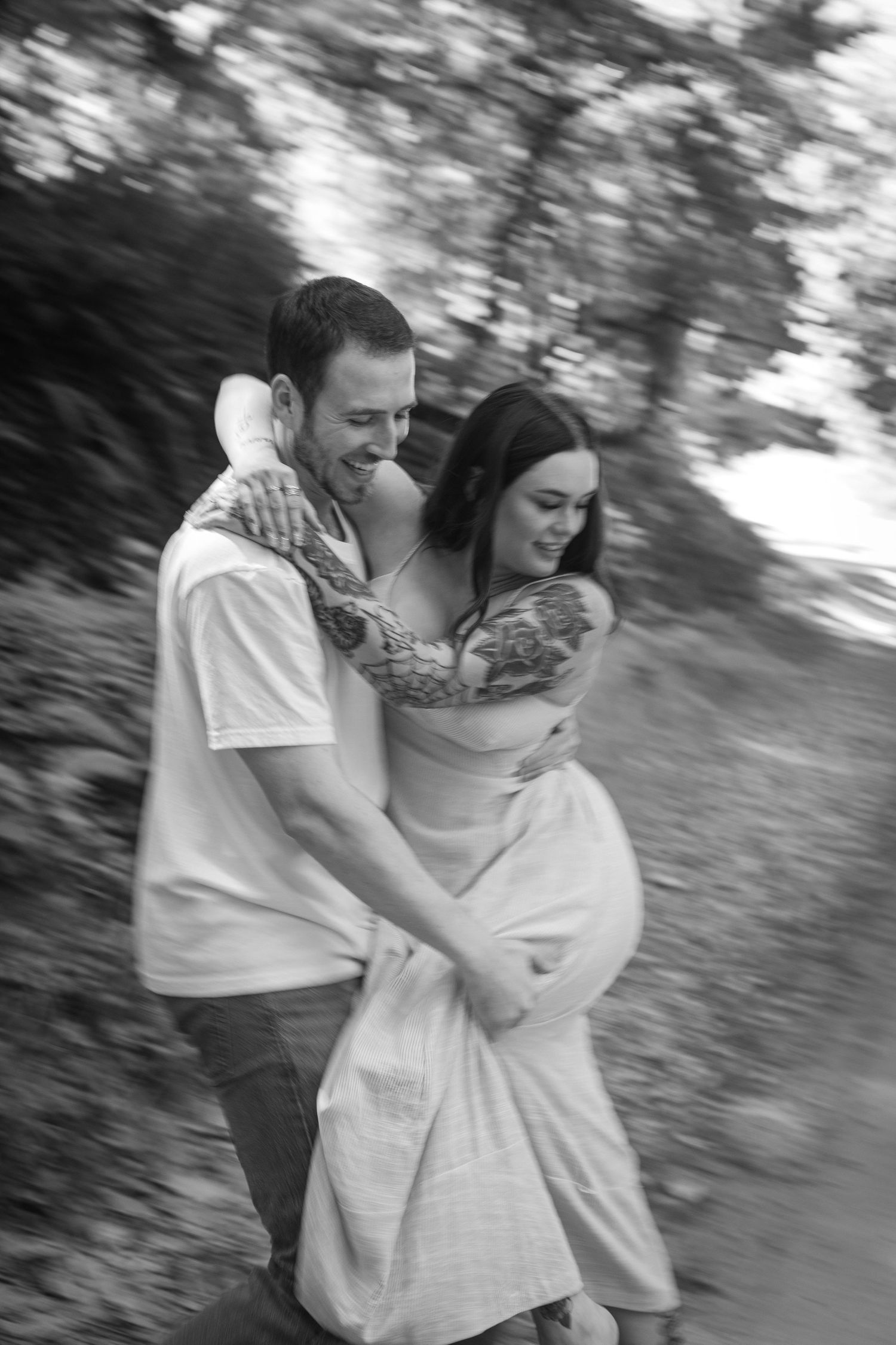 Blurred motion black and white photo of couple dancing and laughing on forest path.