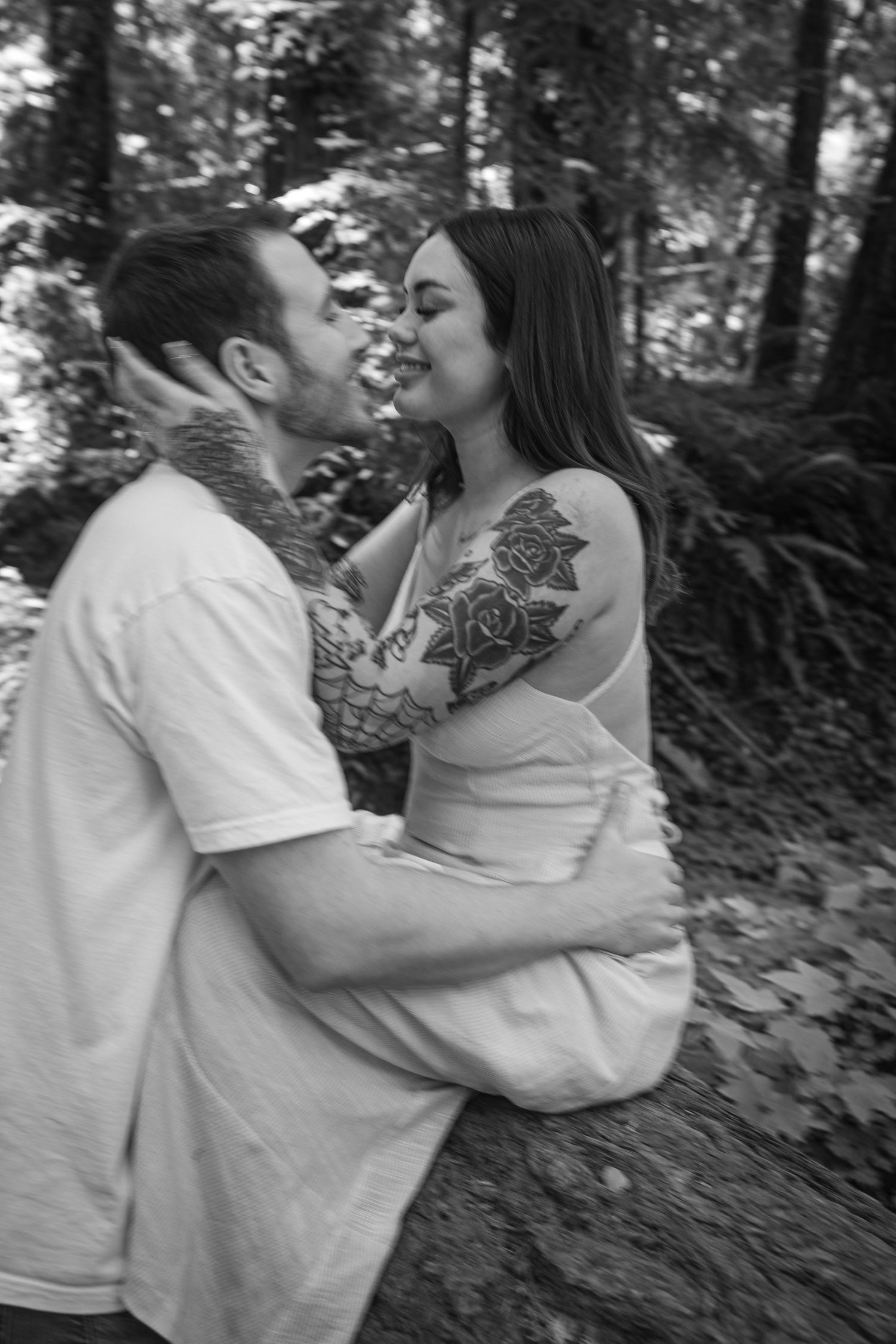 Intimate black and white photo of couple embracing on forest trail with visible tattoos.