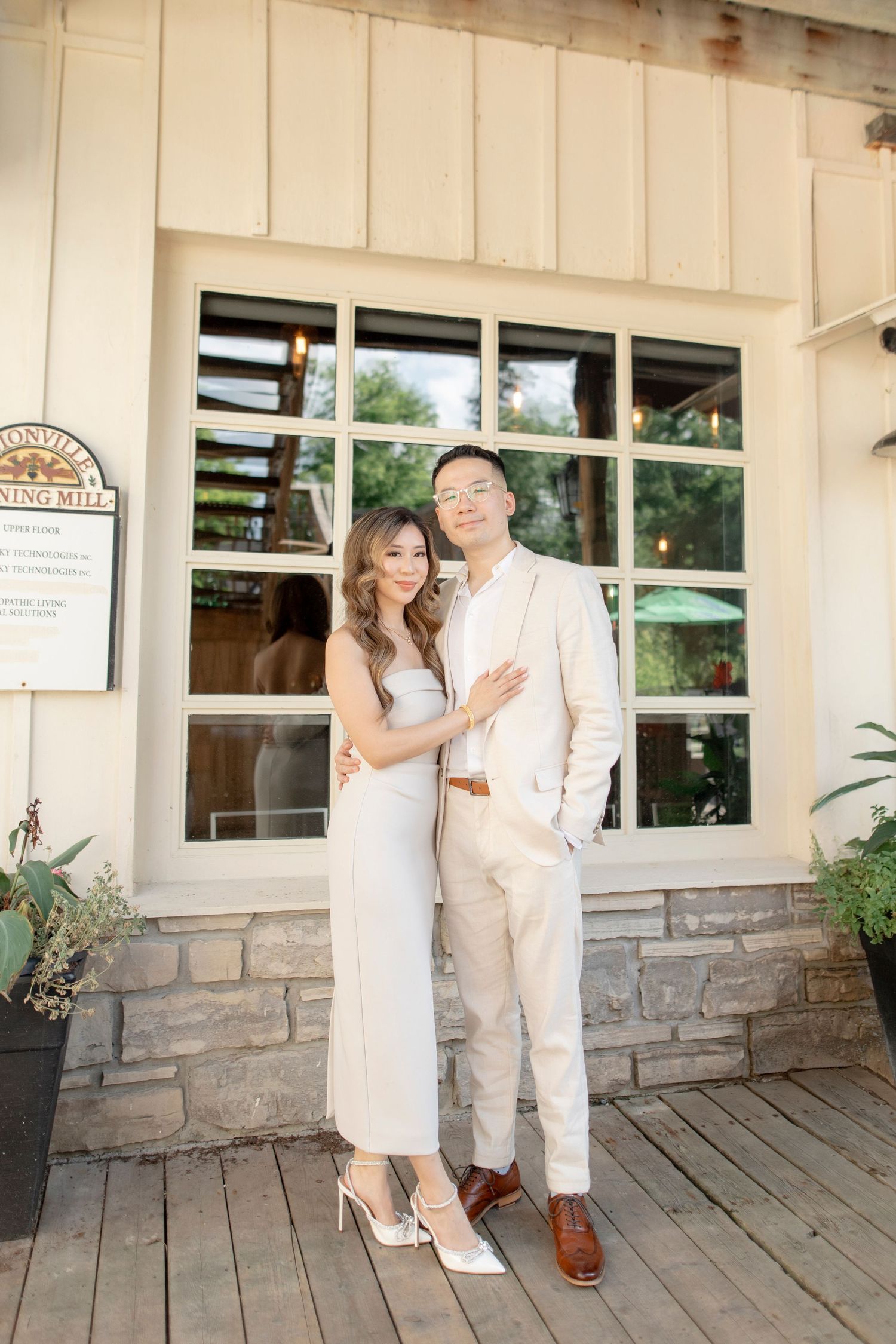Couple poses in coordinated white outfits outside a rustic building with stone and wood details.