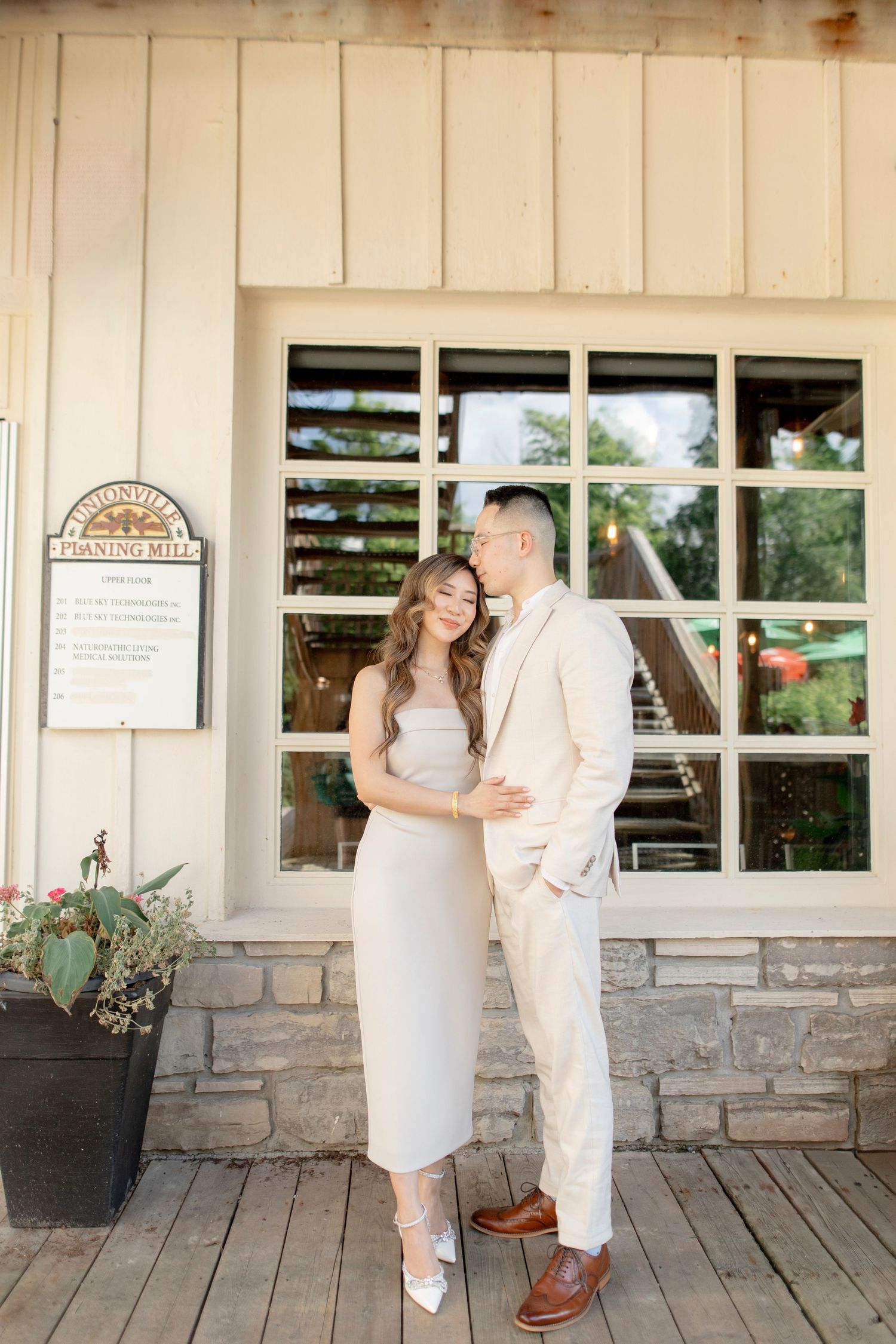 Couple embraces in front of a cream-colored building with stone foundation and large windows.