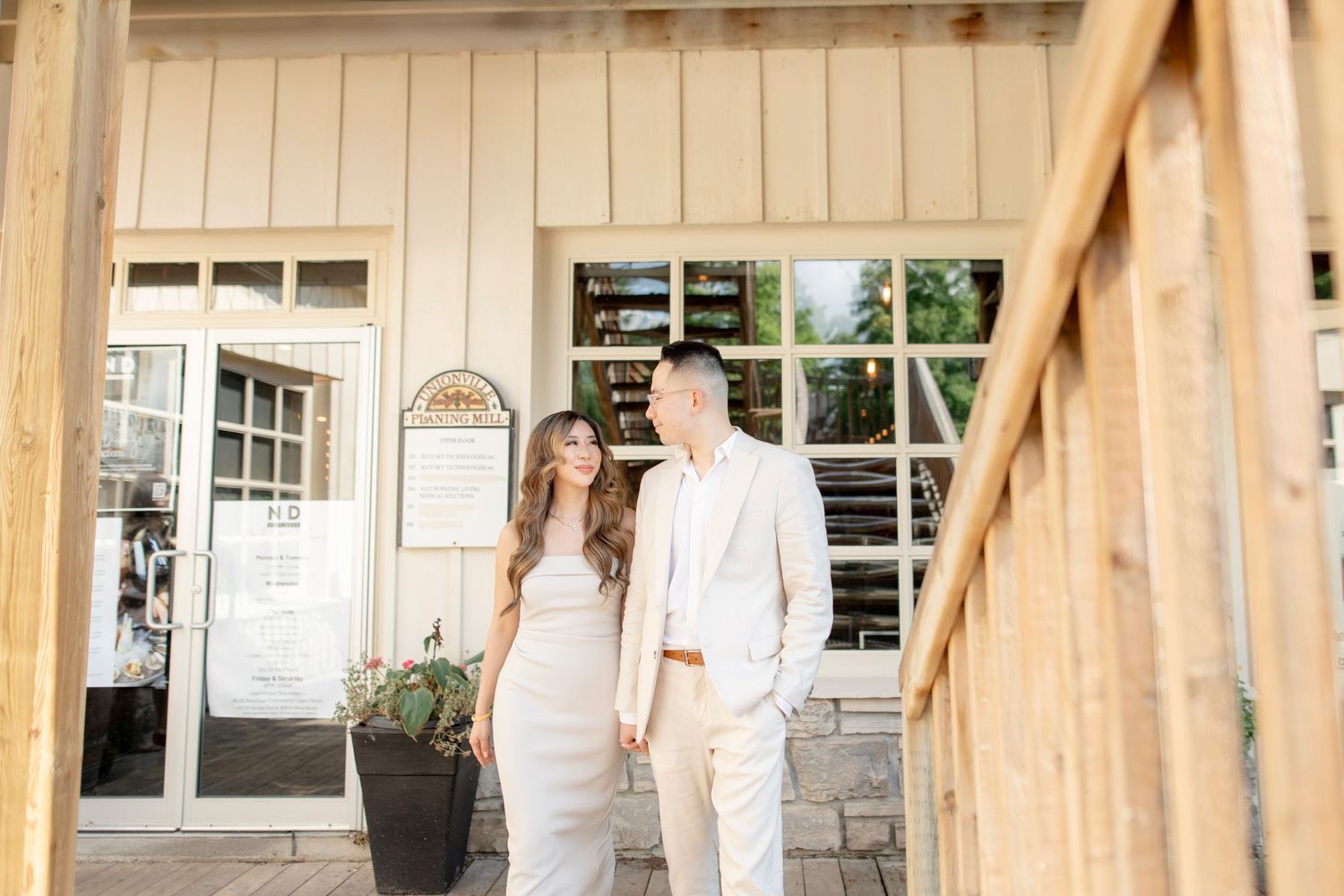 Couple in coordinated white attire stands on wooden deck outside rustic building entrance.