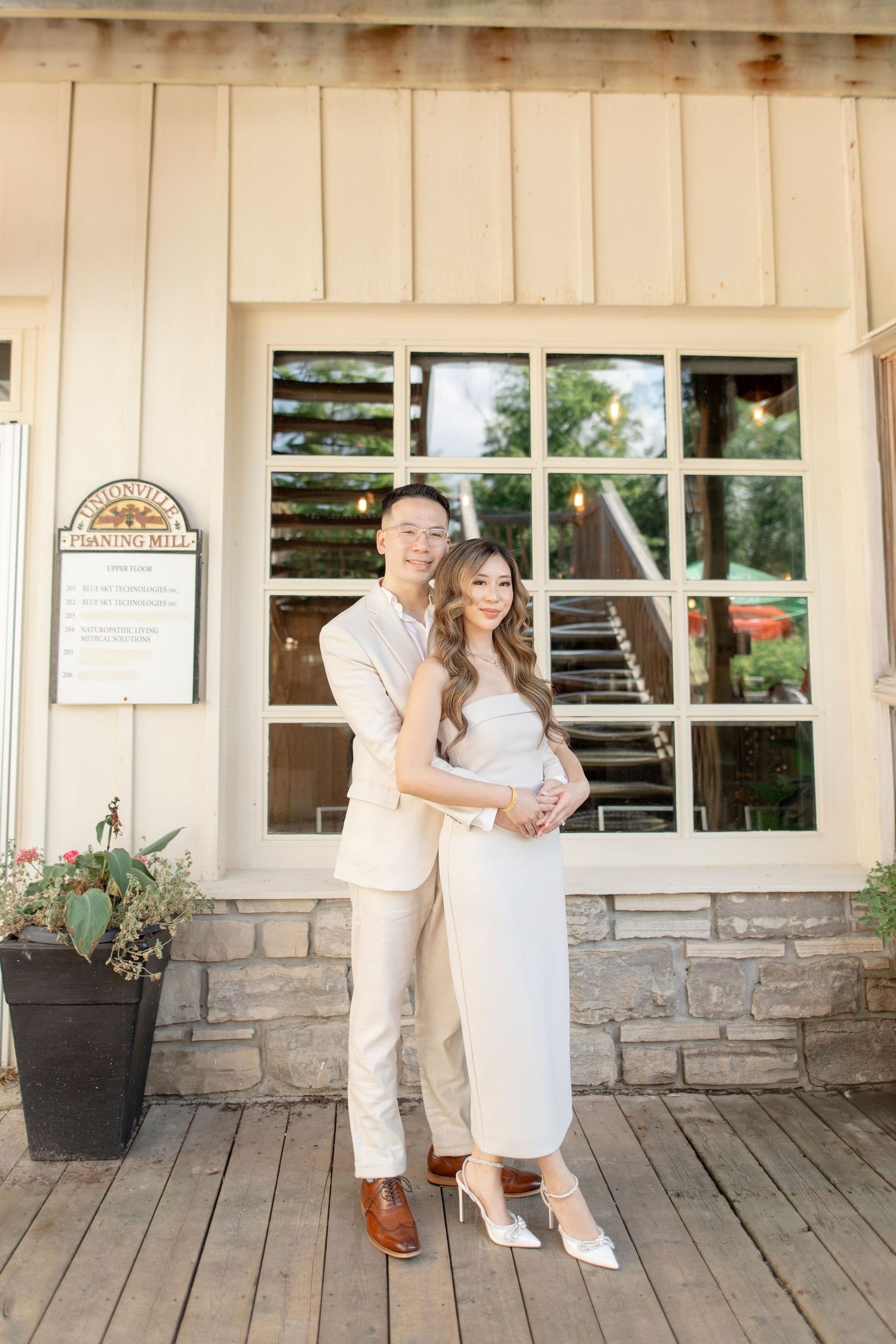 A romantic couple in coordinated cream outfits embrace on a rustic wooden porch with stone accents.