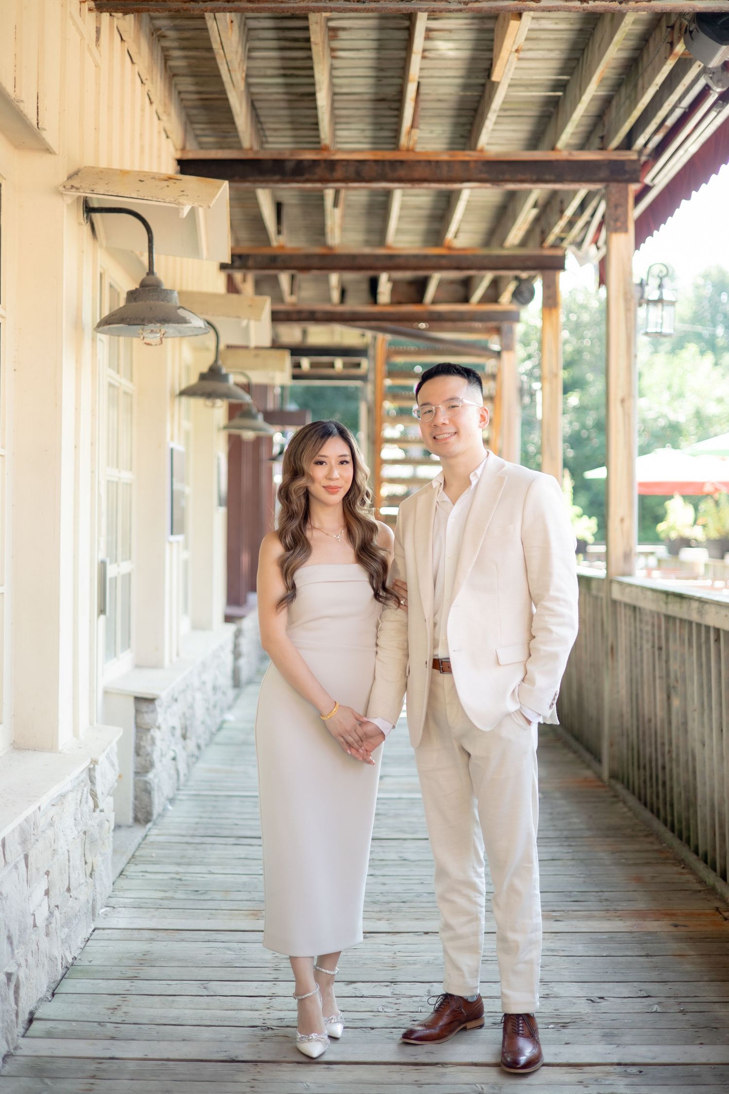 A couple in matching cream formal wear pose on a covered wooden walkway with vintage lighting.