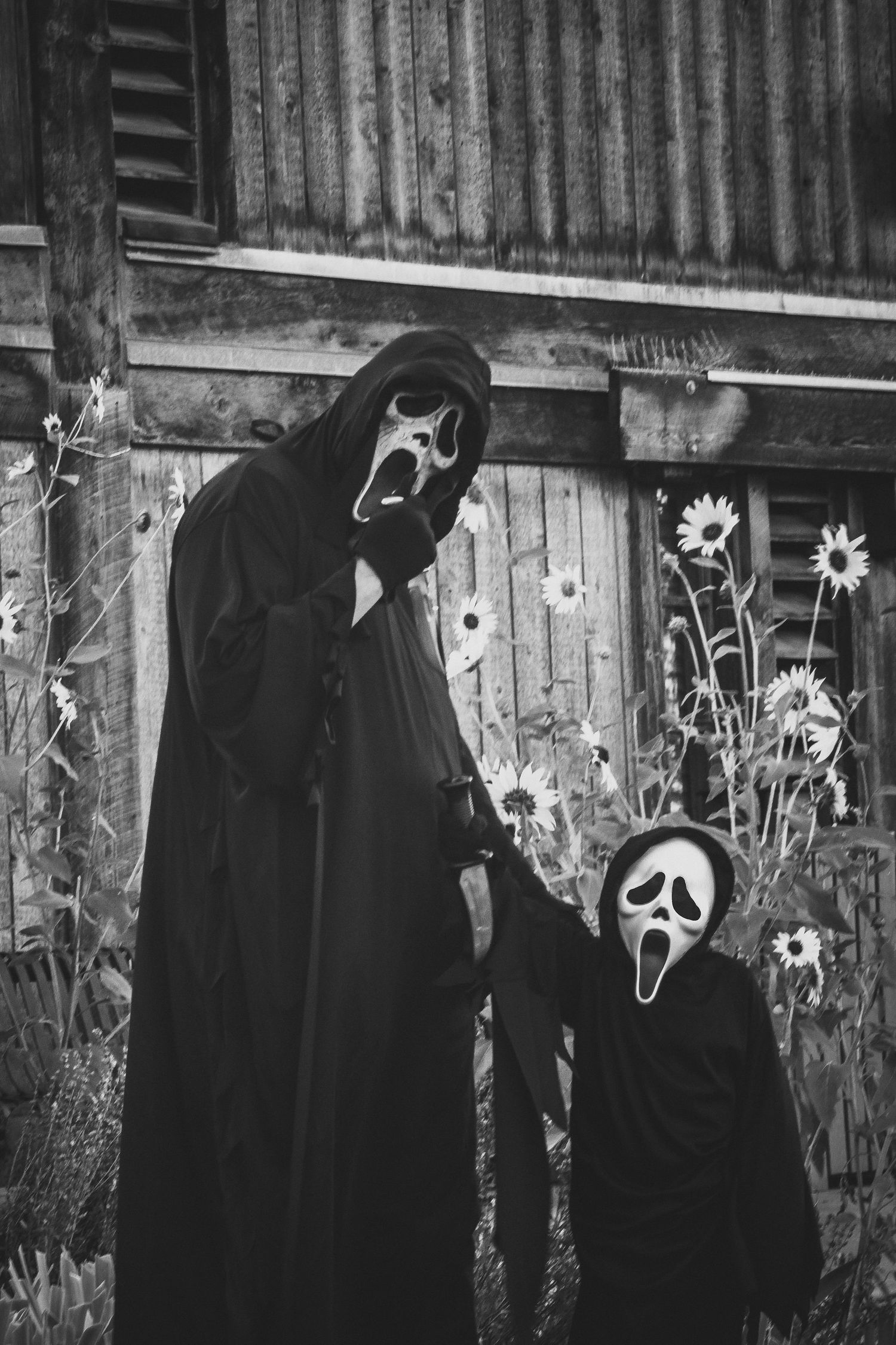 A tall and small figure in Ghostface masks pose together by a wooden wall with flowers.