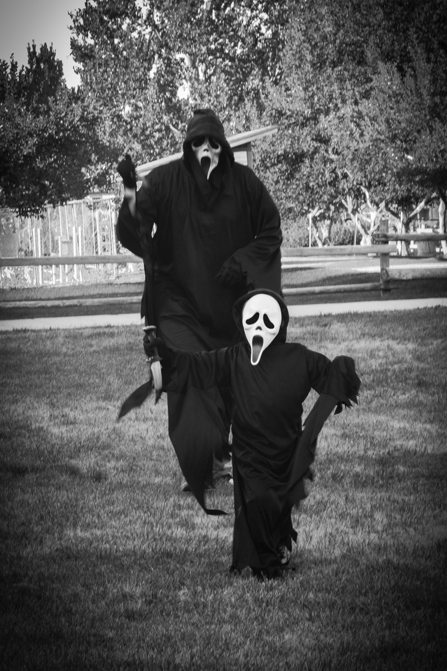 Two people in Ghostface costumes pose on a lawn in black and white.