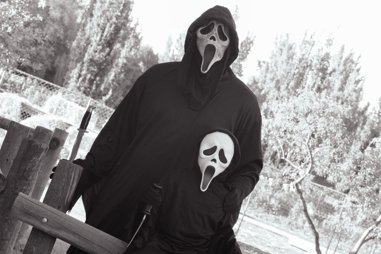 A person in Ghostface mask and dark costume stands outdoors in monochrome.
