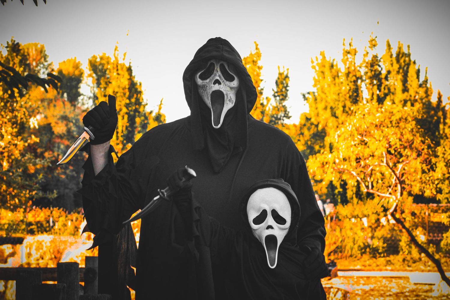 Two figures in Ghostface costumes pose against autumn trees with orange foliage.