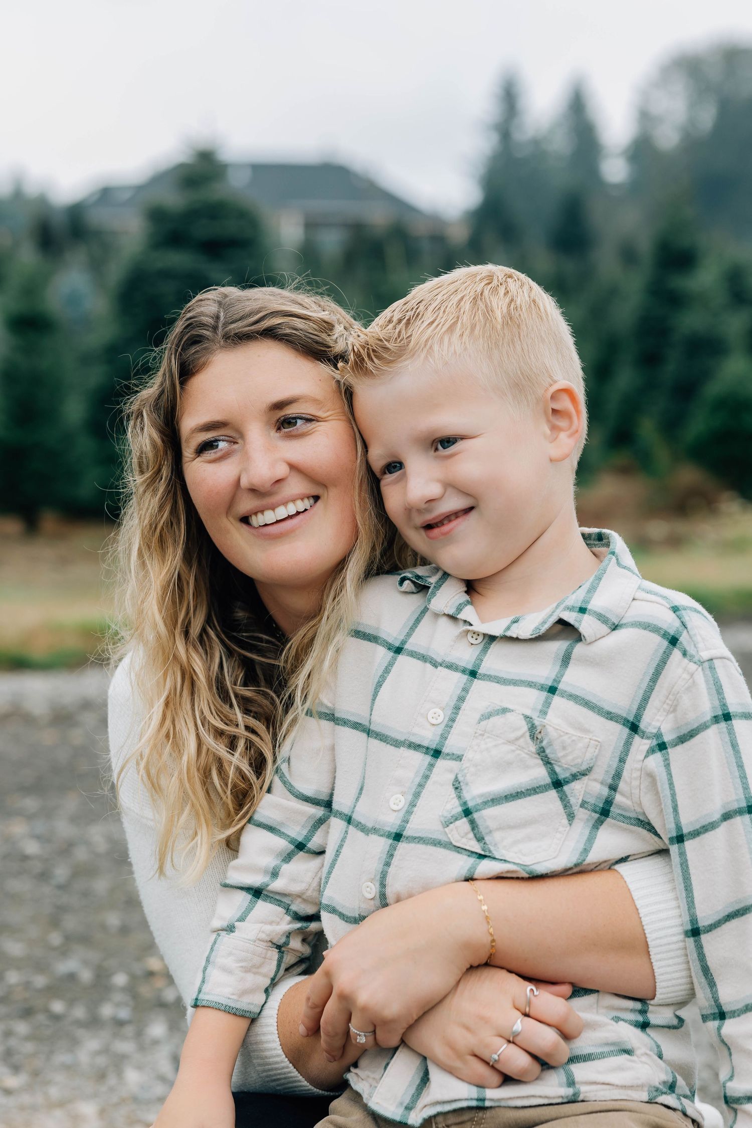 Smiling mother with long blonde hair hugging her young son at a Christmas tree farm.