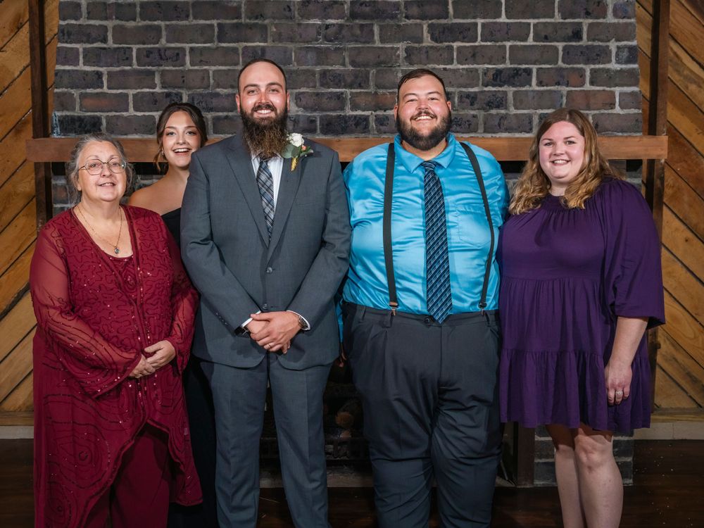Wedding party group portrait wearing formal attire against a rustic brick and wood backdrop.