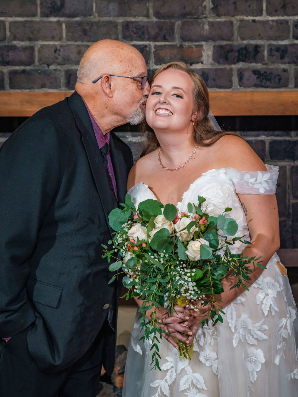A tender moment shared between a bride in a white lace dress holding a green and white bouquet against a brick wall.