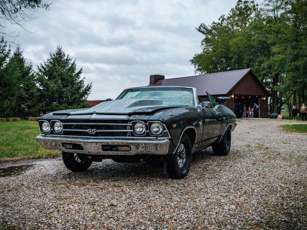 Classic black Chevelle muscle car parked on gravel path in front of rustic wooden building.