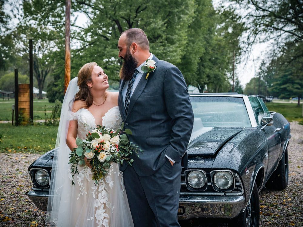 Newlyweds share a moment beside a classic black vintage car during their outdoor wedding.
