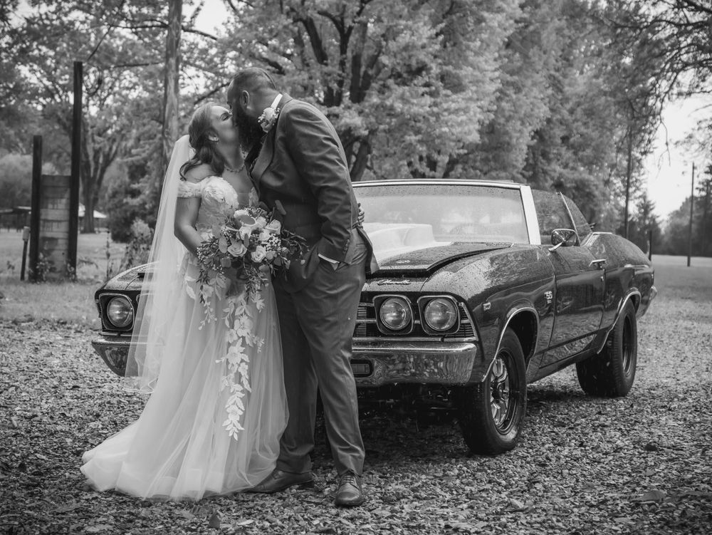 Black and white portrait of newlyweds with a vintage Chevelle car in a wooded setting.