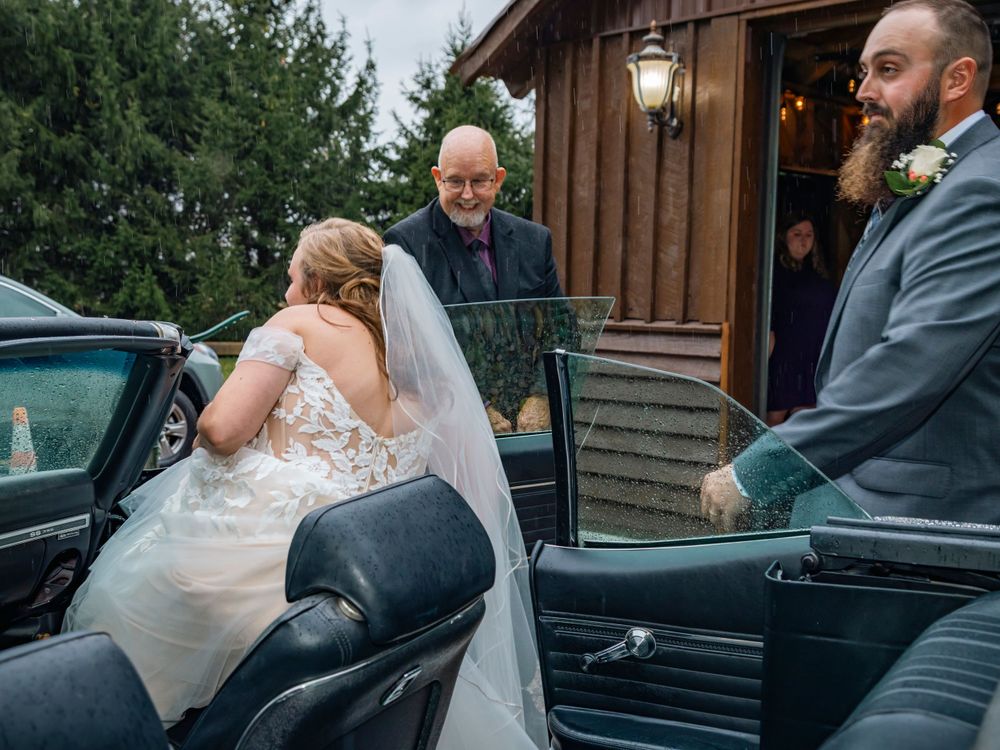 Wedding couple getting into a classic car outside a rustic wooden venue.