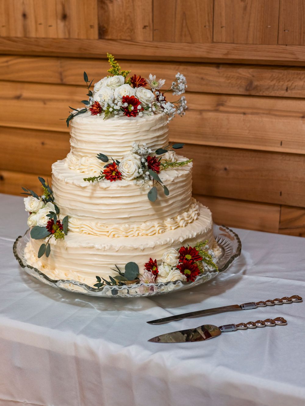 A textured white wedding cake with burgundy flowers and greenery on a white tablecloth.