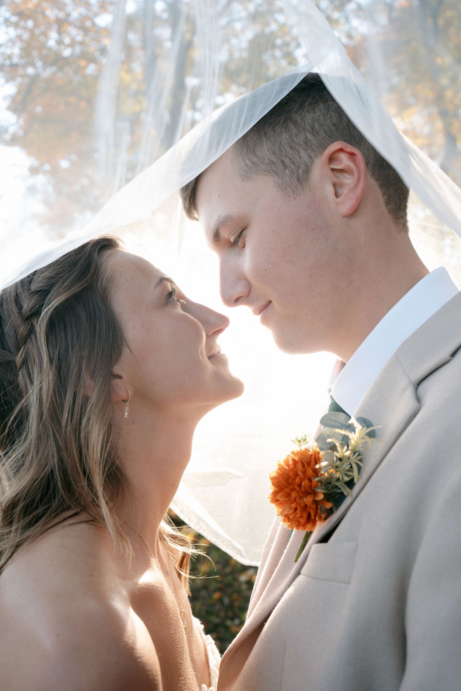 Bride and groom sharing romantic moment under wedding veil with soft backlit autumn trees behind them.