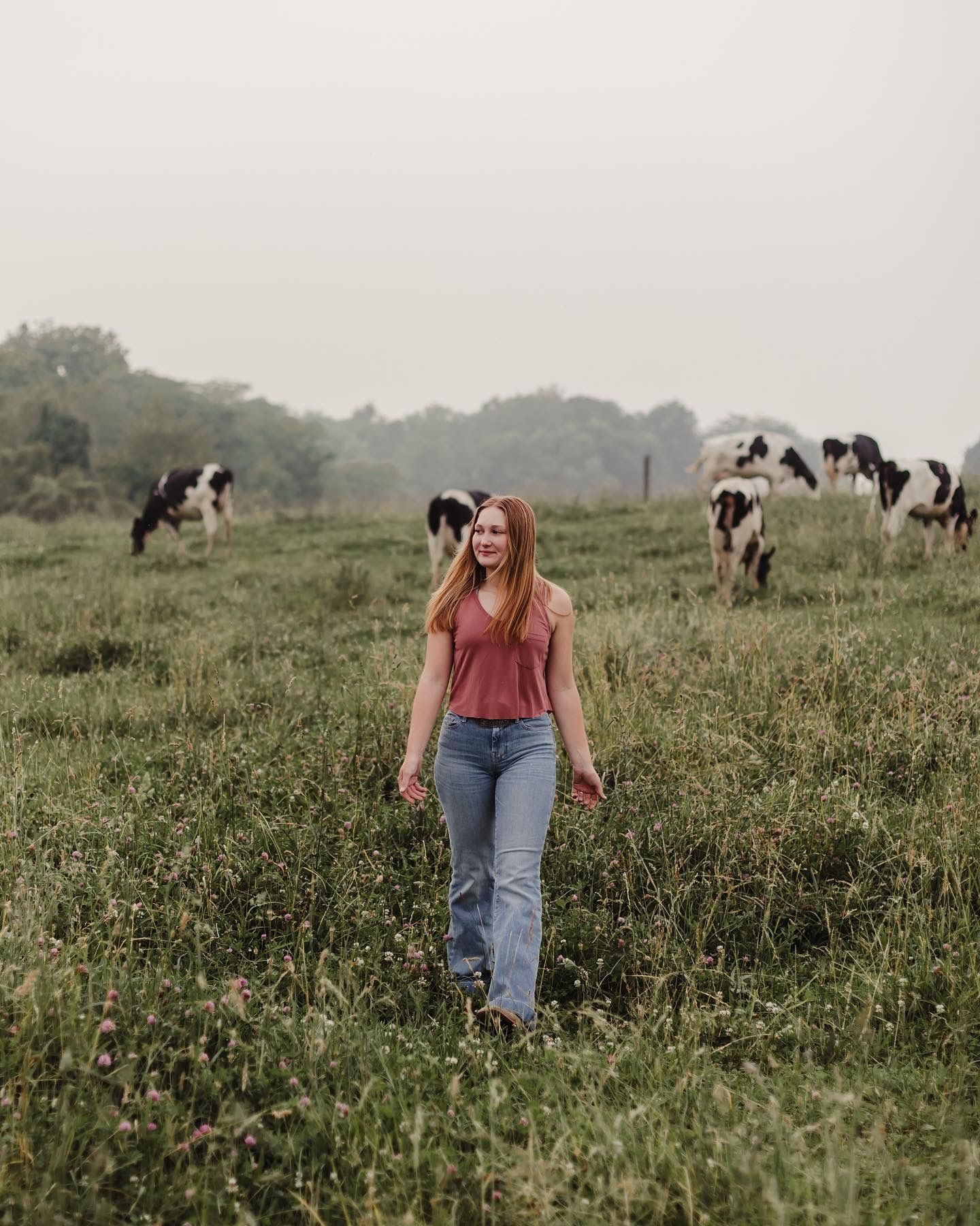 Young woman in red tank top and jeans smiling while walking through a lush green field with cows in background.