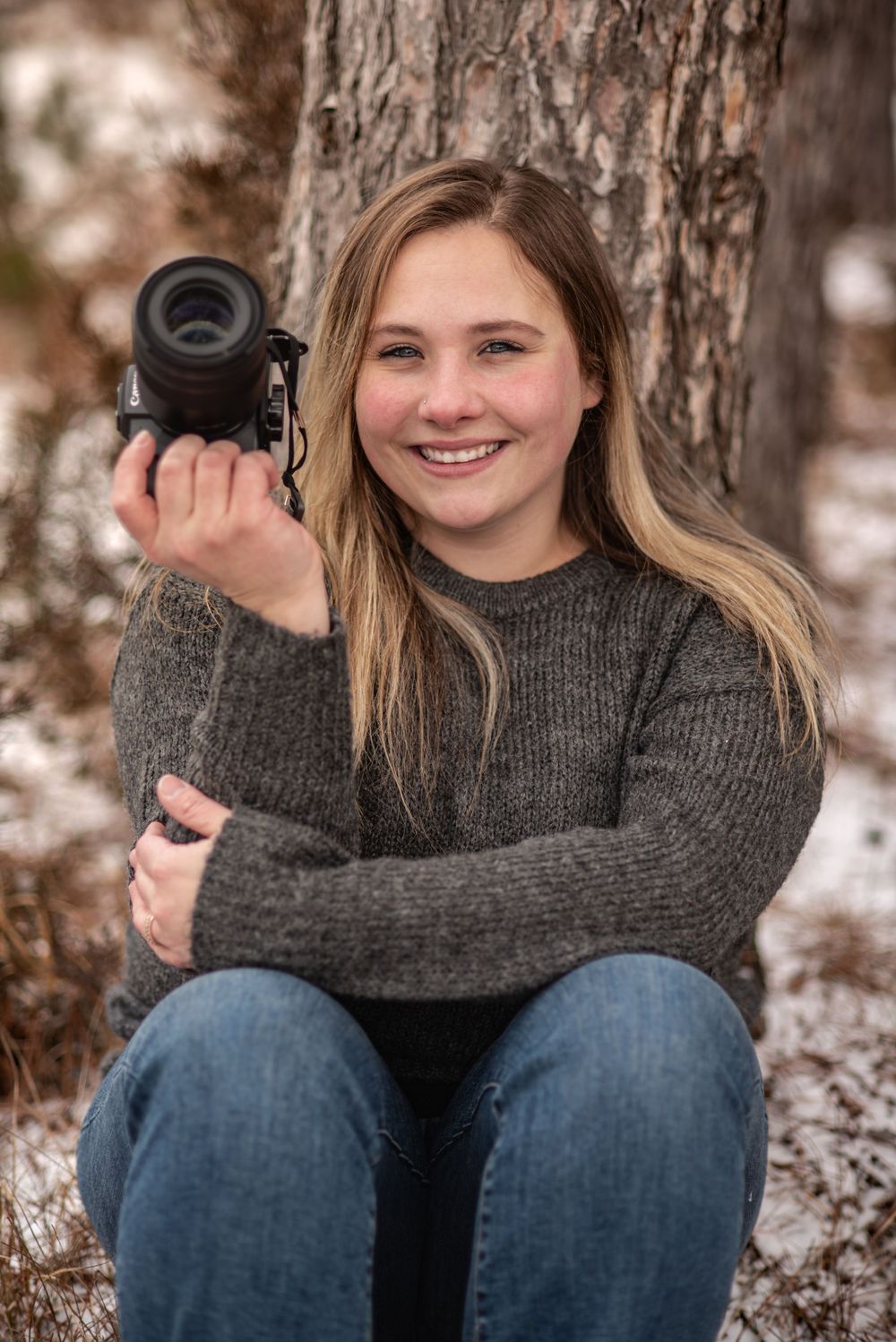 Young photographer in gray sweater holding camera while sitting against tree trunk in winter setting.