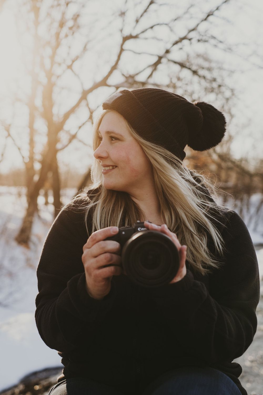 Person in winter clothing holding camera outdoors during golden hour.