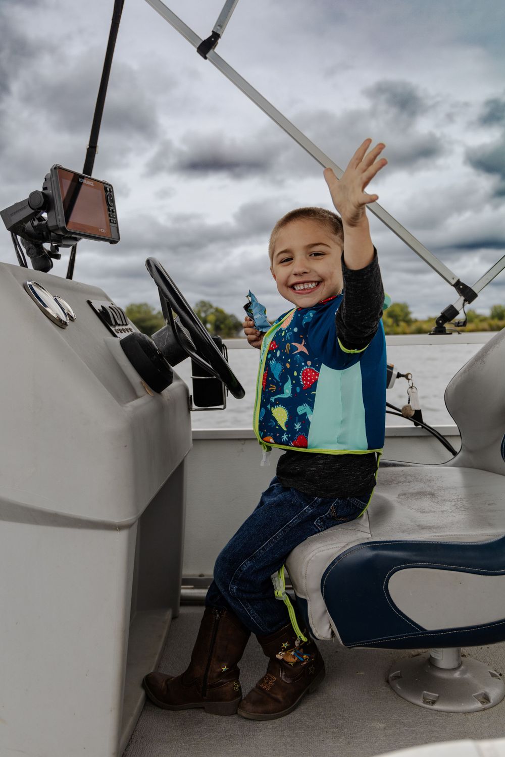 Young passenger waves excitedly while seated at a boat's control panel.
