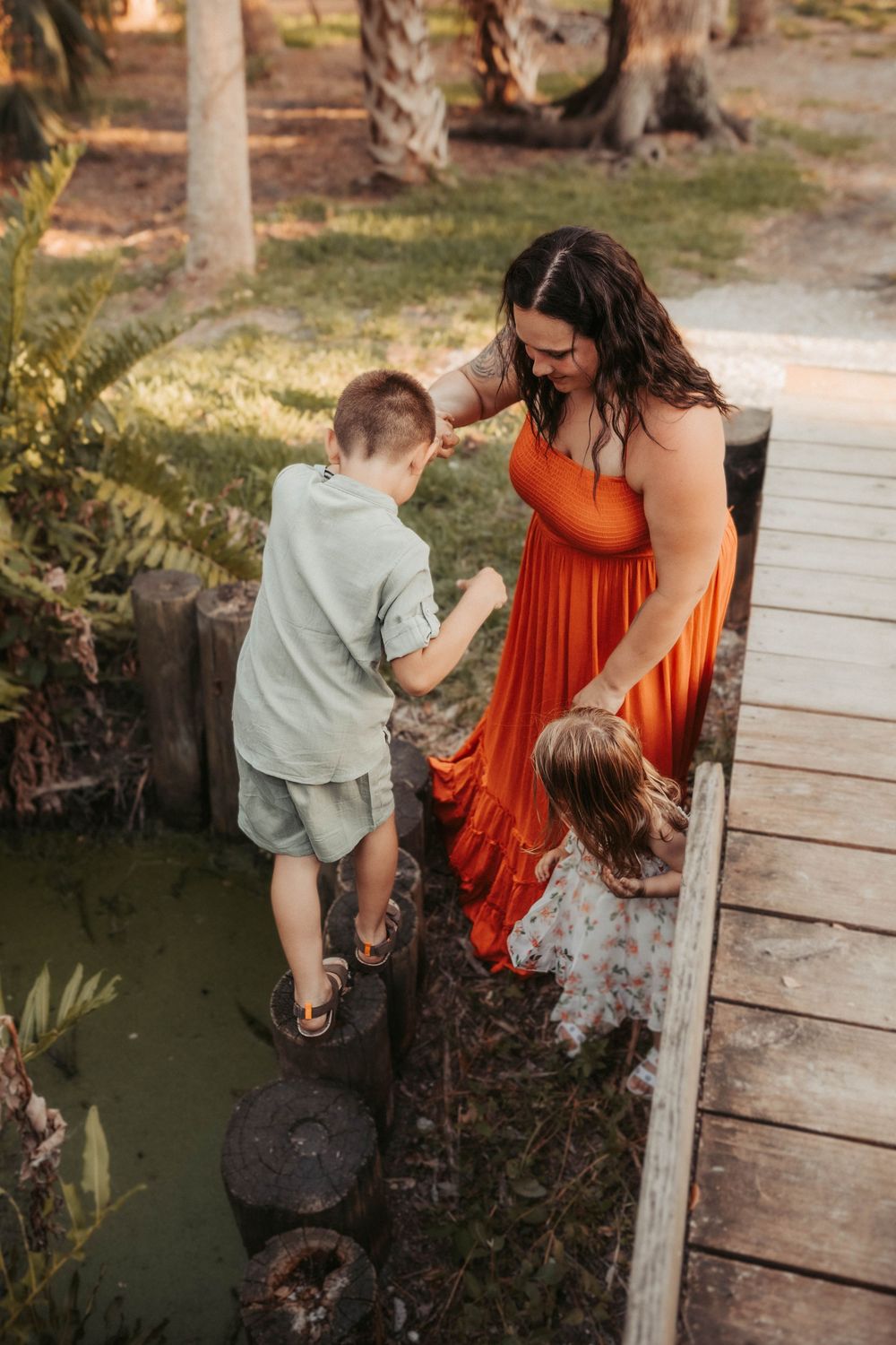 A woman in an orange dress stands on a wooden boardwalk with two children near water and palm trees.