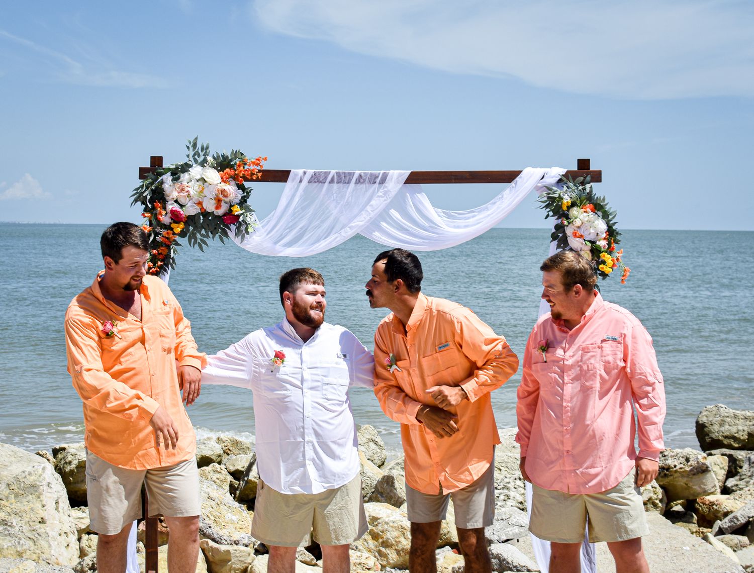 Groomsmen in coral and pink shirts chat under a floral wedding arch by the ocean.
