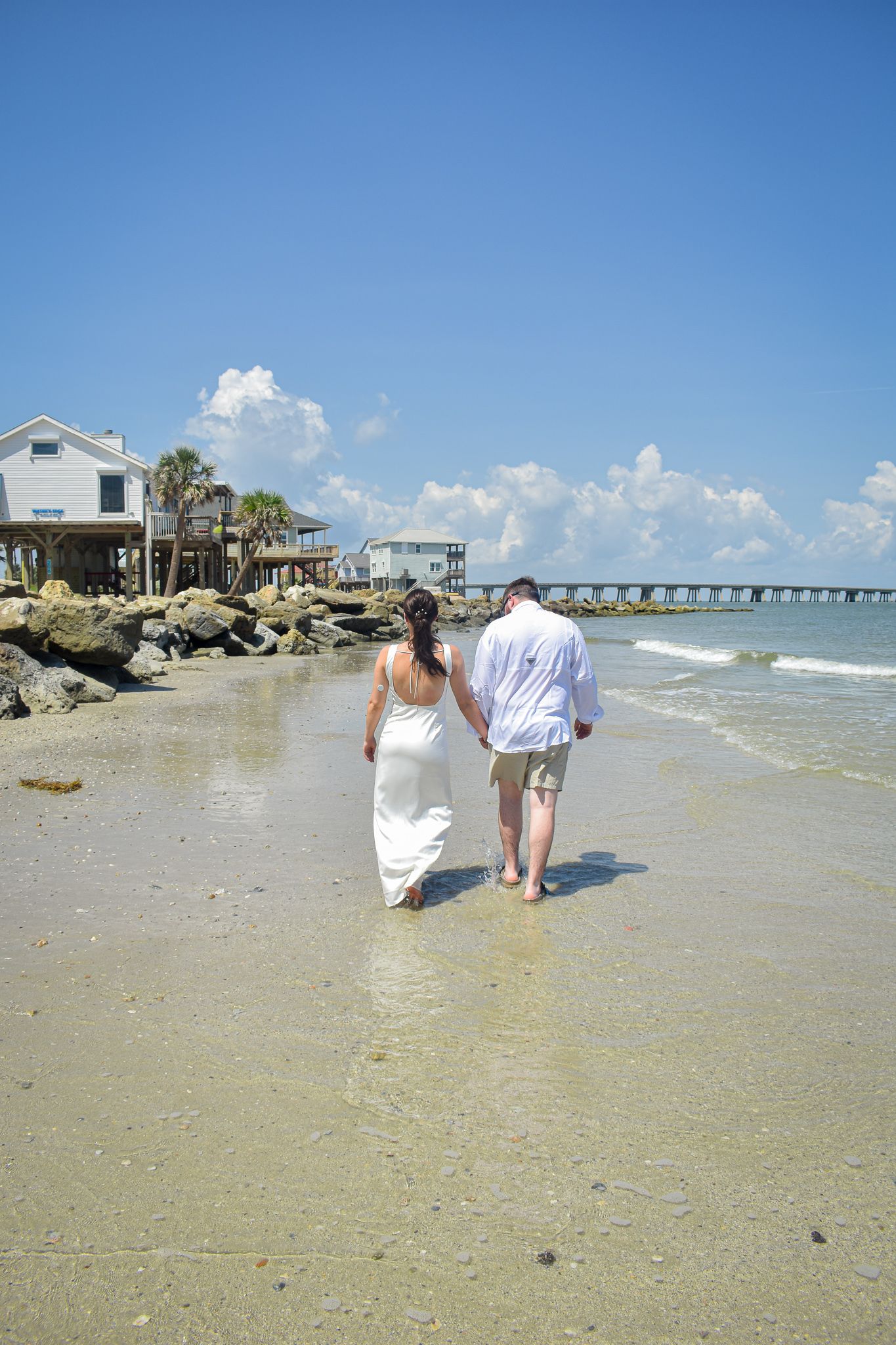 Couple walking along a sandy beach with beach houses and pier in the background on a sunny day.
