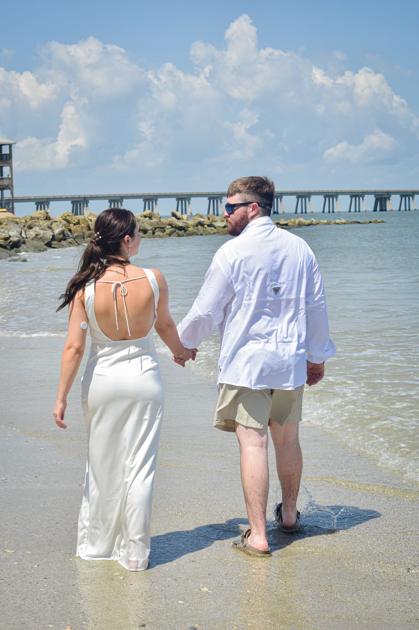 Couple walking hand in hand along a beach with a pier in the background.