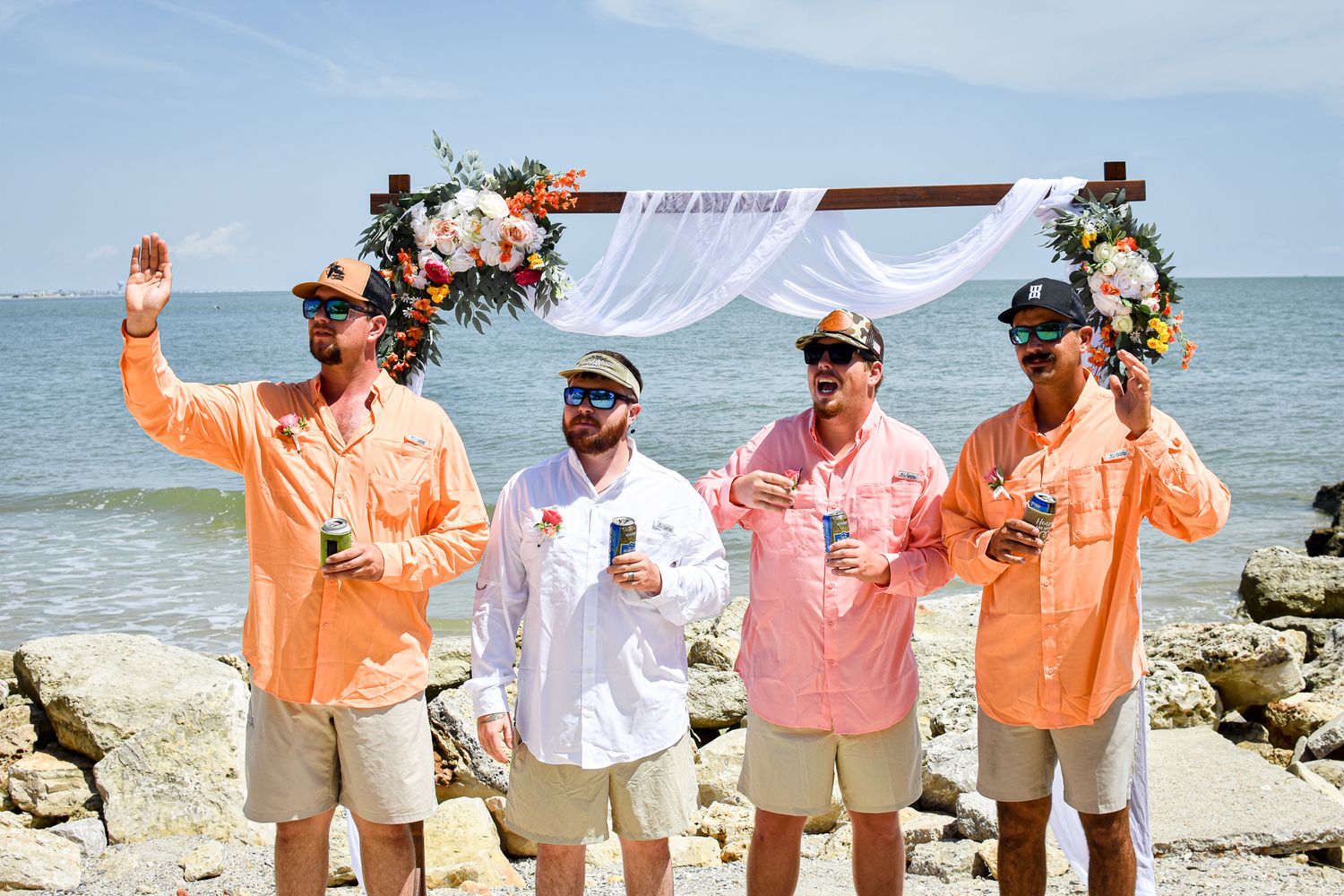 Wedding party in orange and pink shirts celebrating on a rocky beach with ocean views.