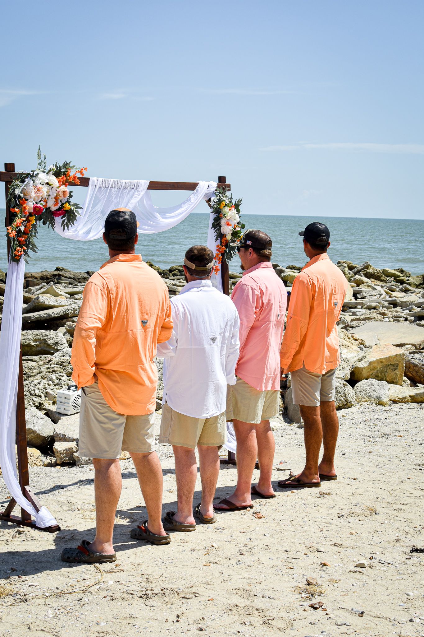 Group in coordinating orange and pink attire standing at beach wedding altar.
