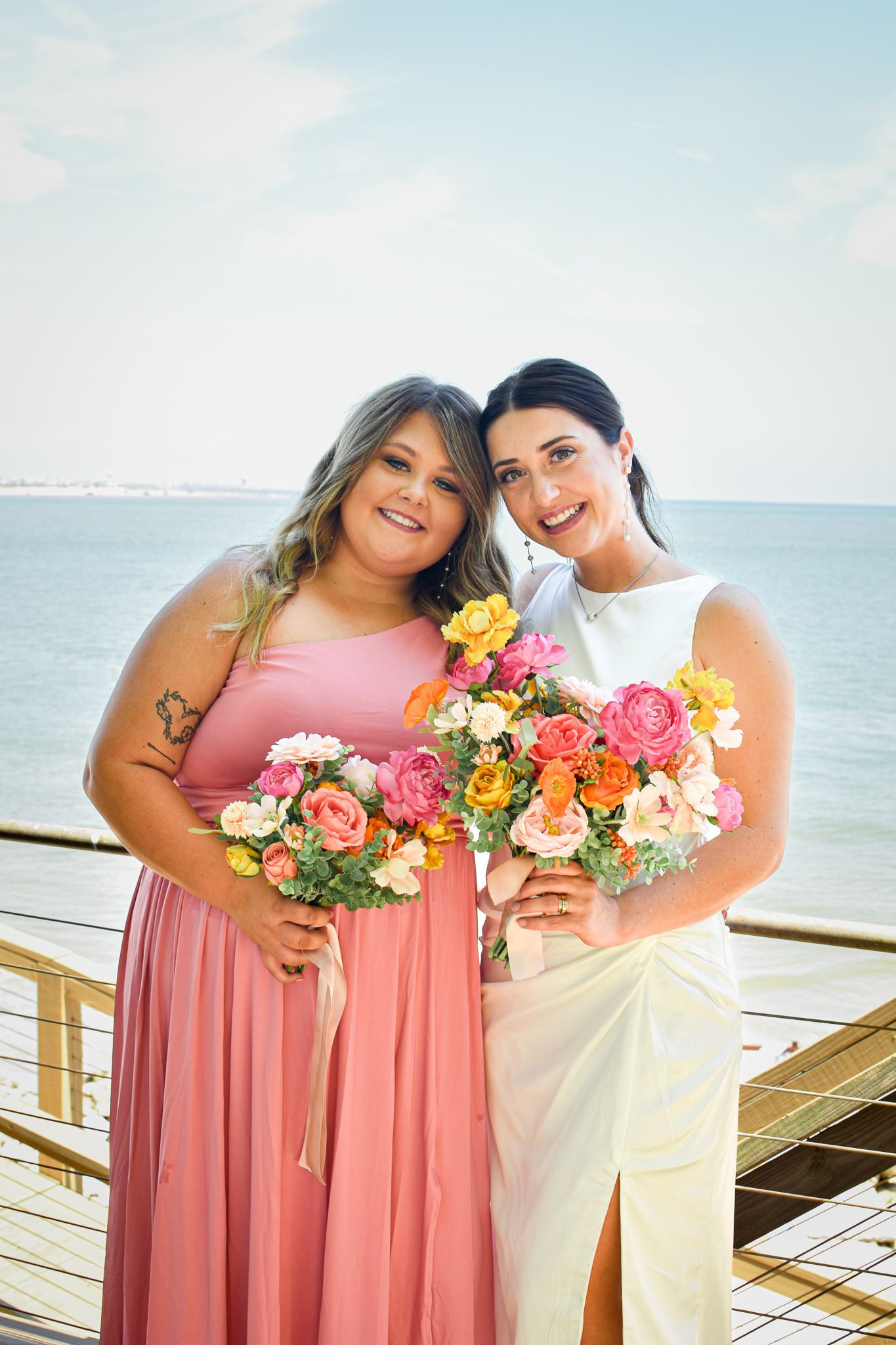 Wedding party members in pink and white dresses holding colorful bouquets on oceanfront deck.