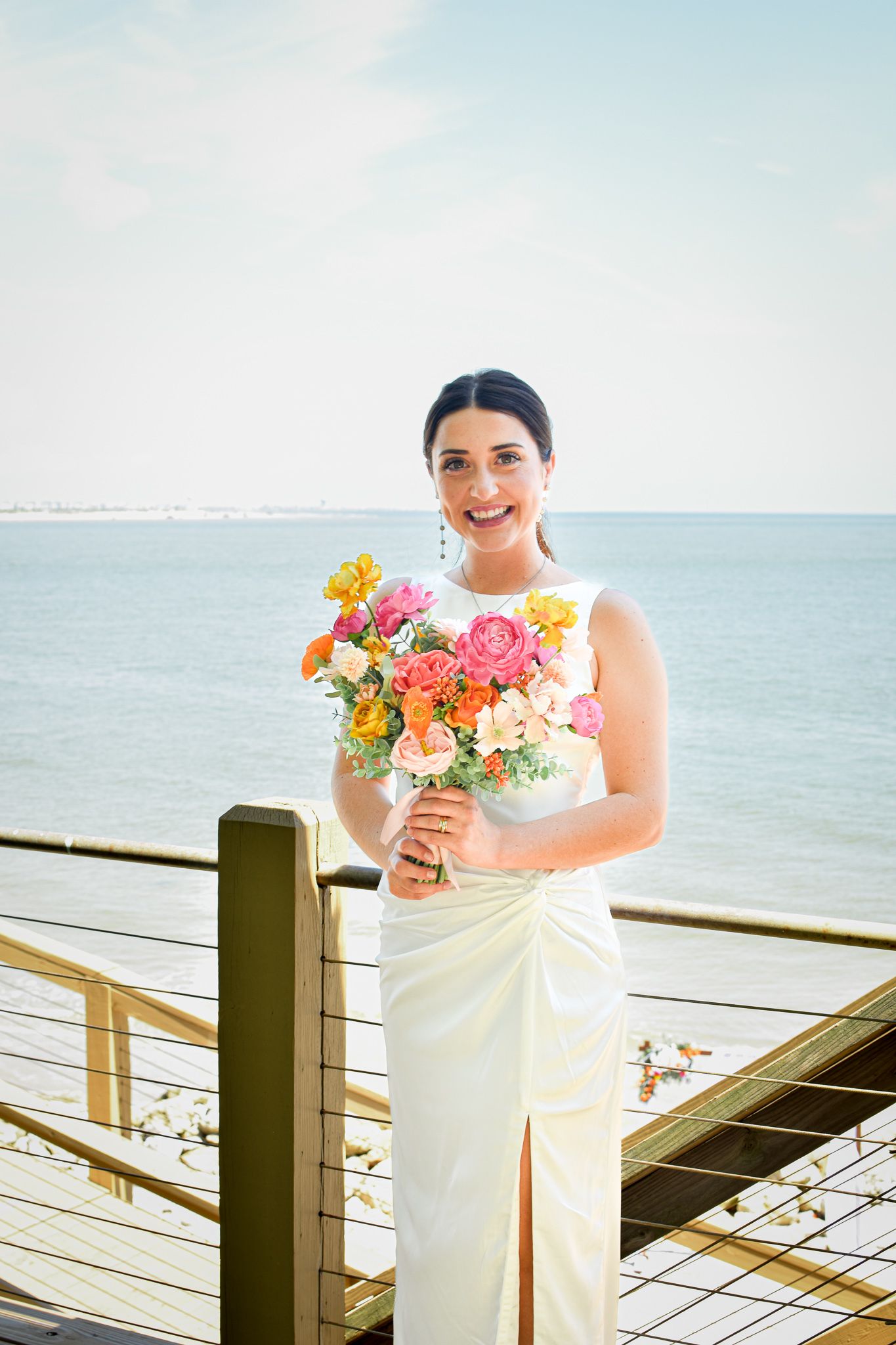 Person in white wedding dress holding vibrant bouquet on oceanfront deck.