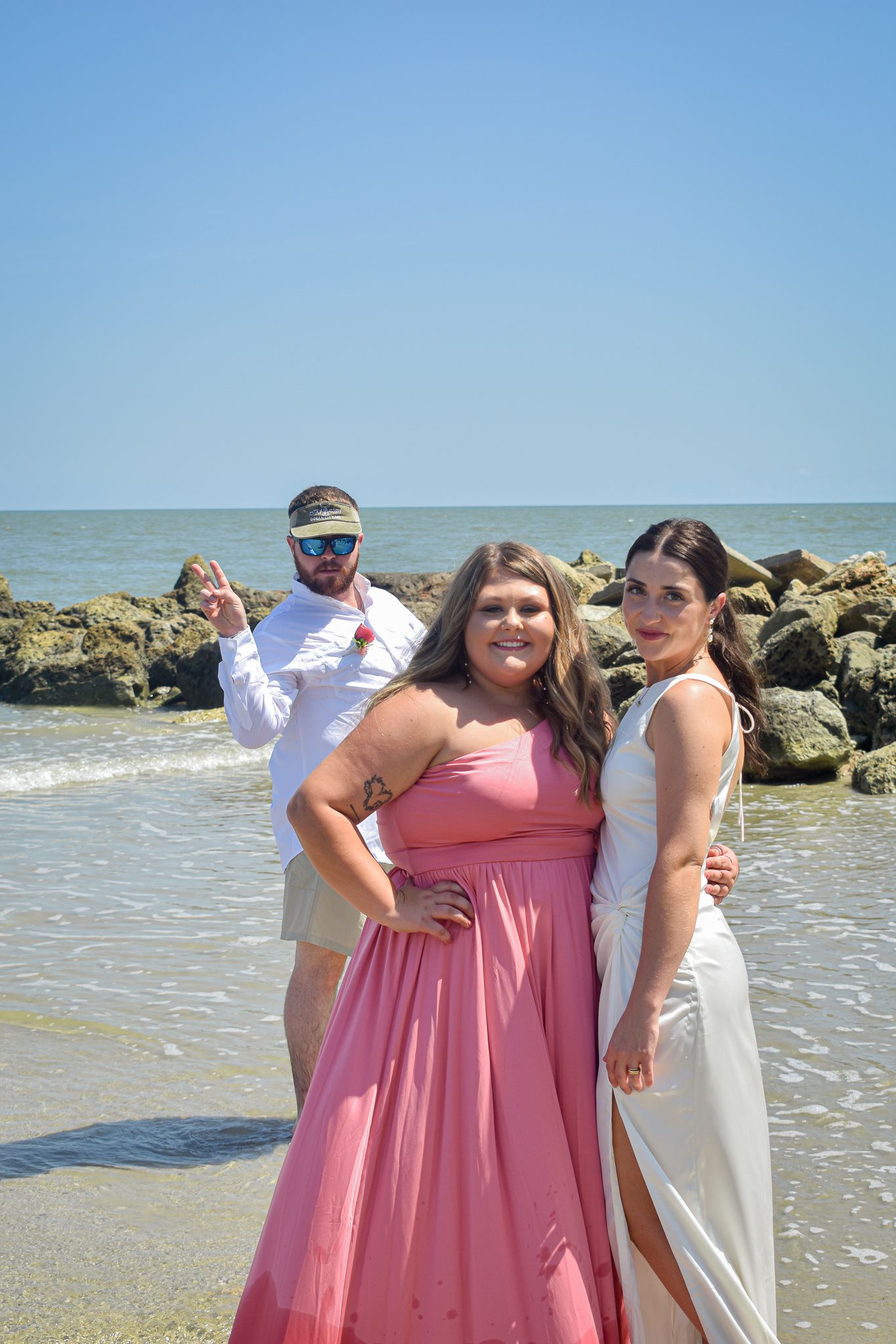 Three people pose together on a sunny beach with rocky coastline in the background.