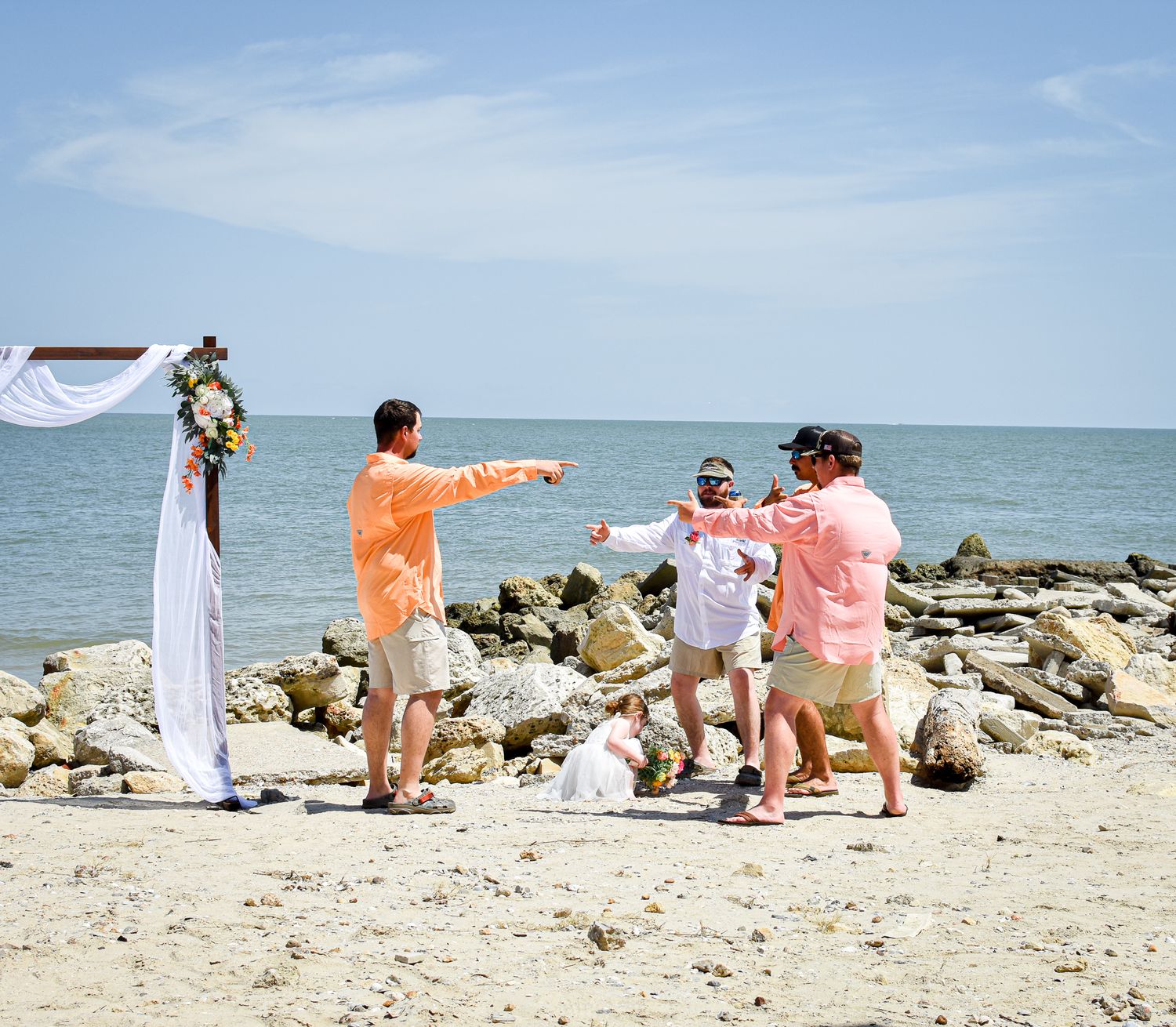 Wedding guests in pastel attire celebrate on a rocky beach under blue skies.