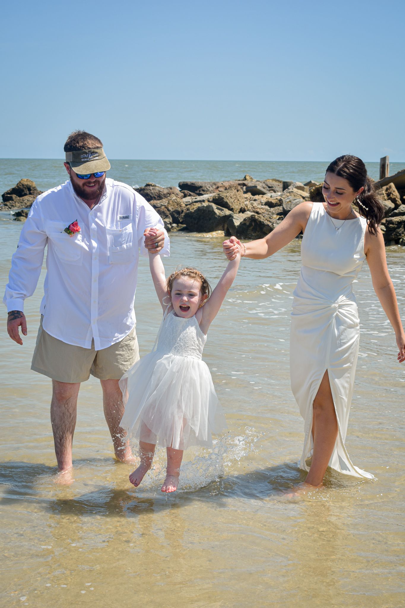 A joyful family moment at the beach as they swing a small child in a white dress between them.