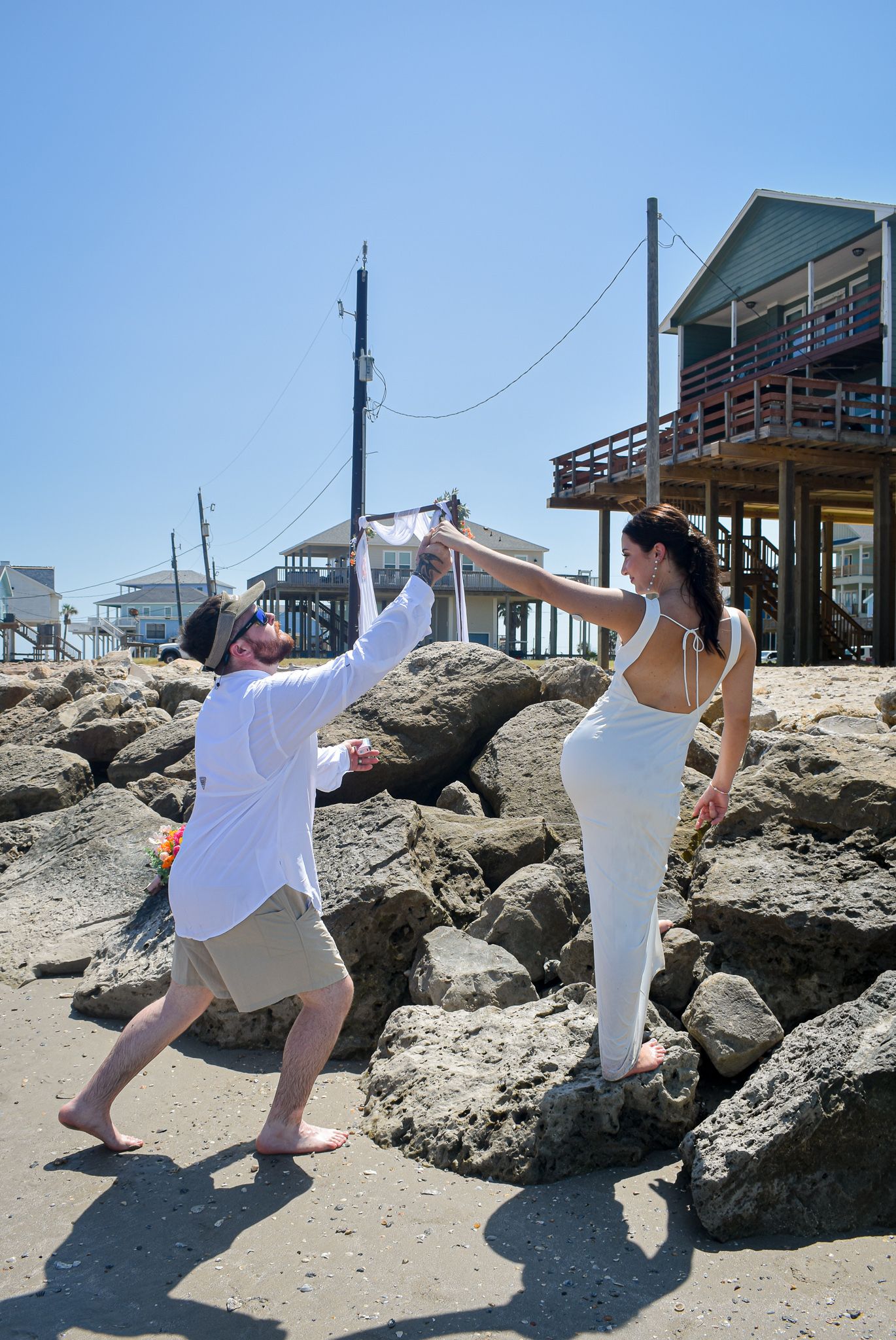 Two people strike playful poses on rocky beach terrain near coastal homes.