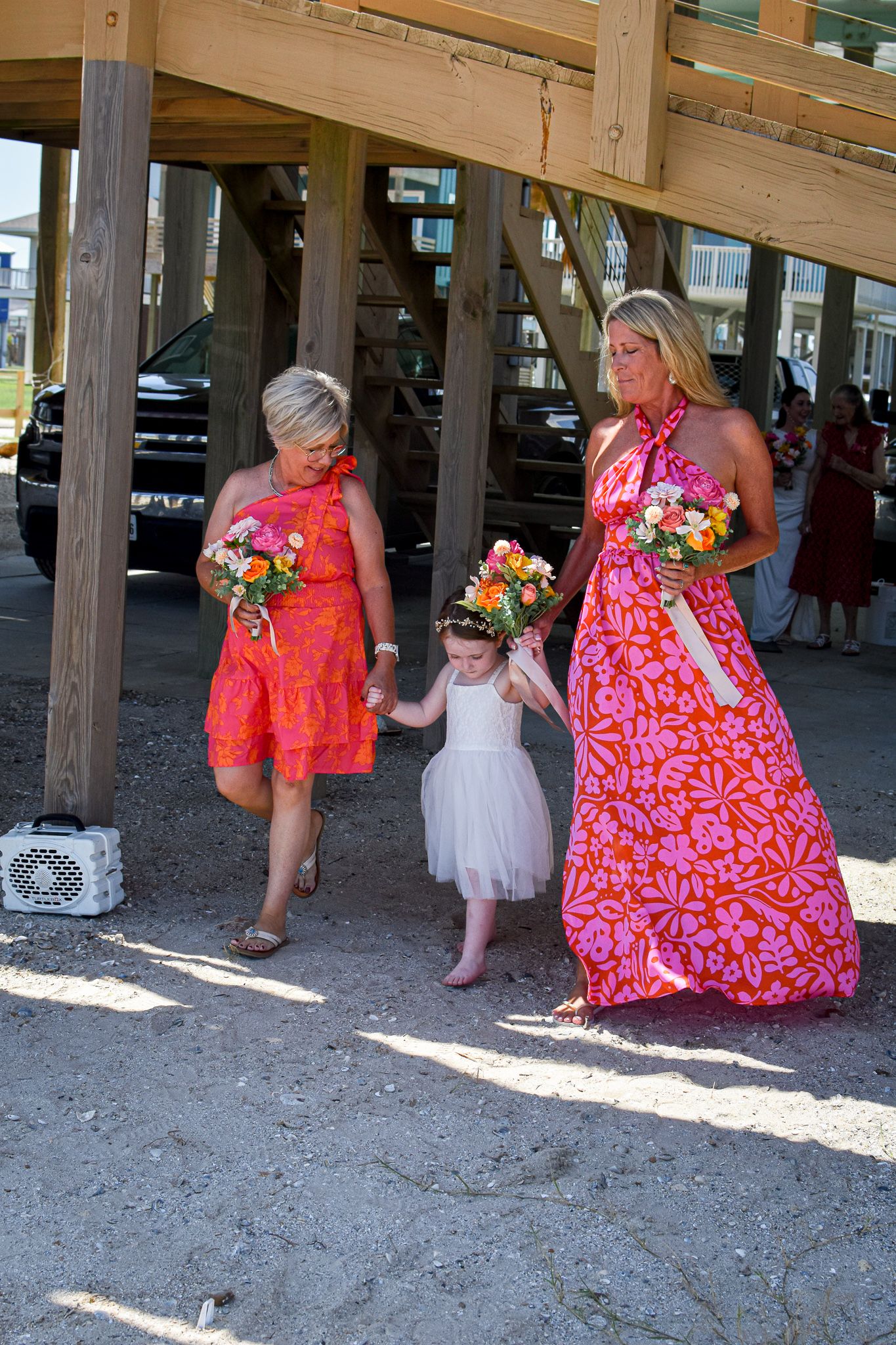 Three generations in bright floral dresses stand under a wooden pier structure.