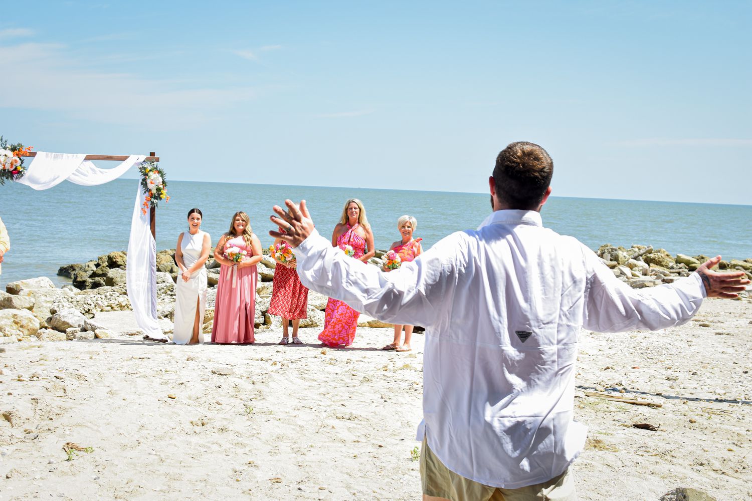 Beach wedding scene with wedding party in coral and white against ocean backdrop.