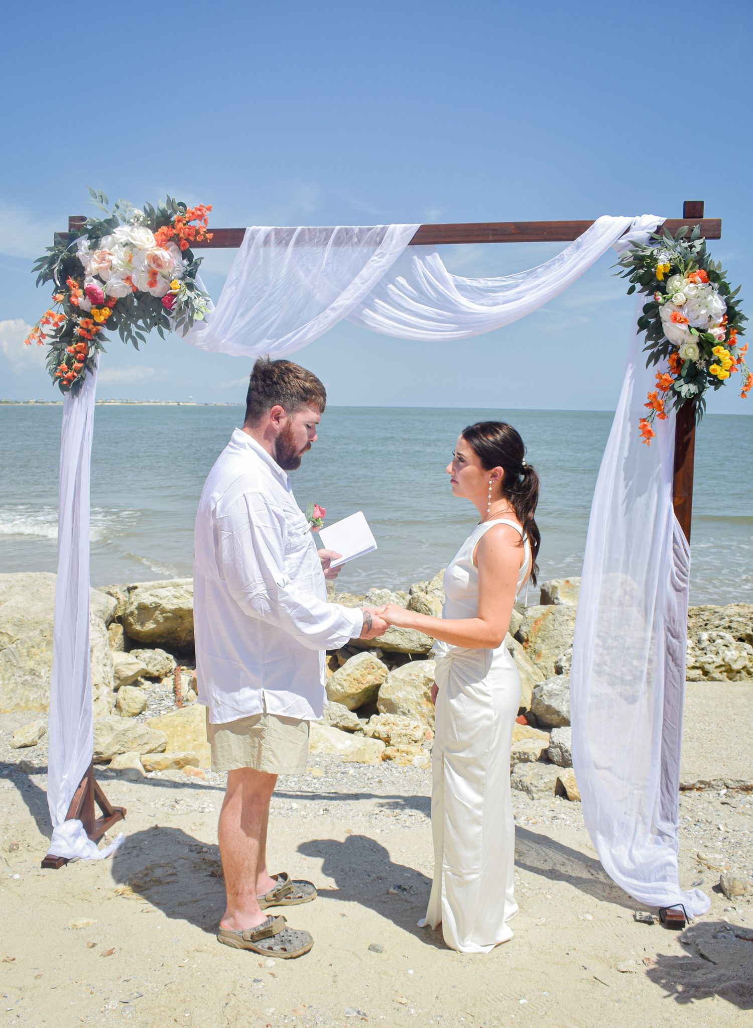 A beach wedding ceremony with a white draped arch decorated with coral and peach floral arrangements by the ocean.